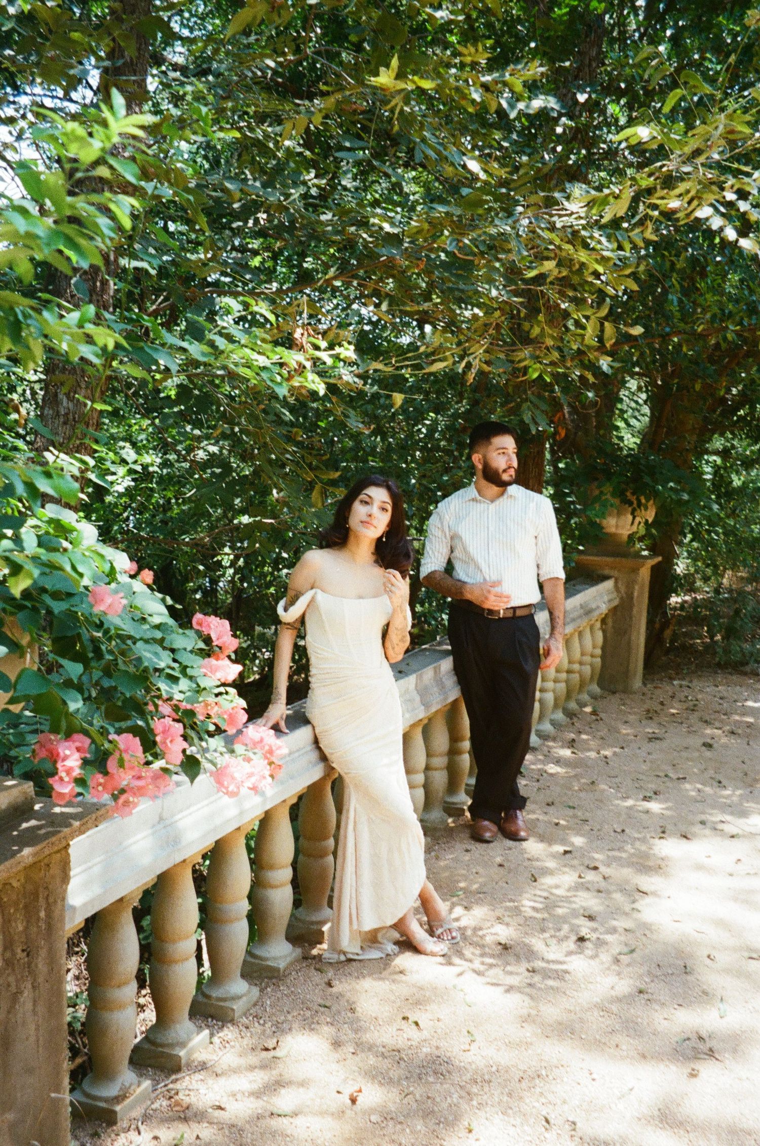 Bride and groom posing on stone balustrade surrounded by lush greenery and pink flowers.