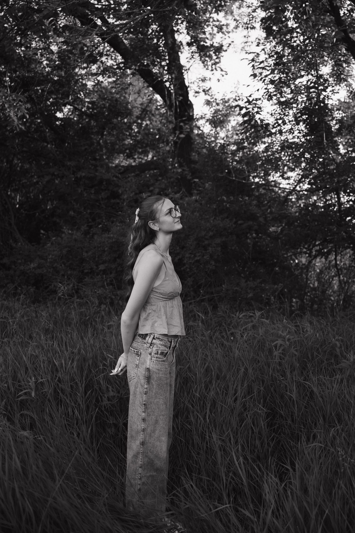 Black and white portrait of woman in tank top and jeans standing in tall grass near trees.