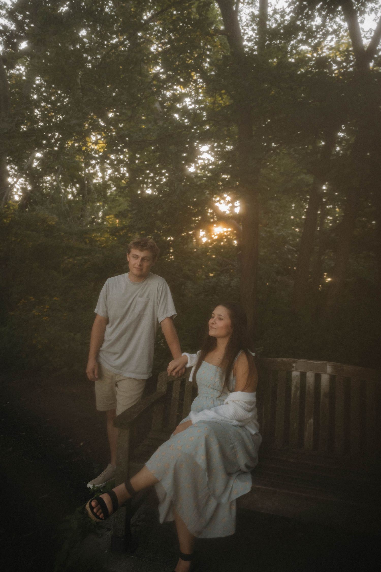 Young couple on wooden deck at sunset, woman in blue dress seated on bench, man standing nearby.