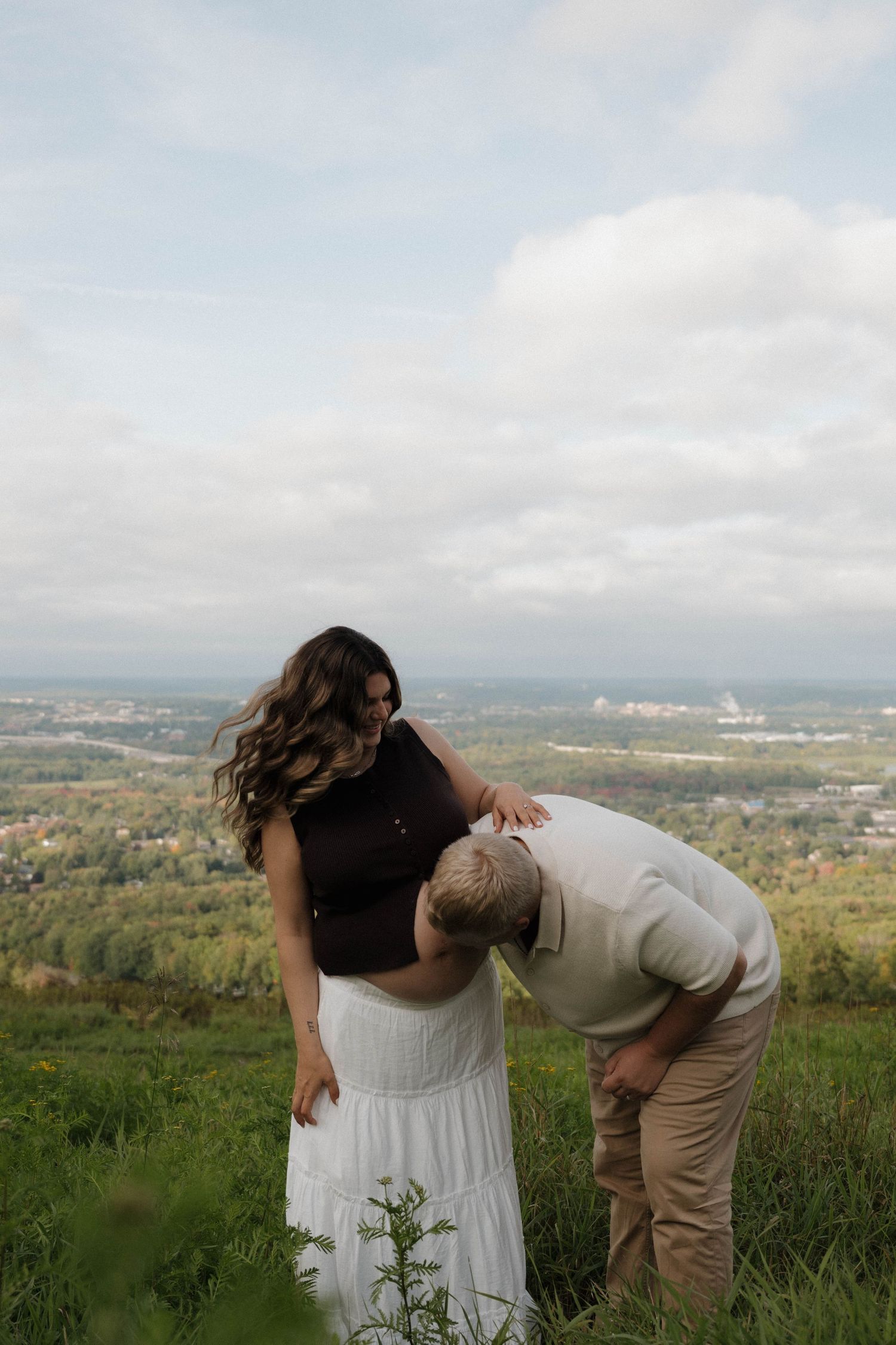 A couple shares a tender moment on a grassy hilltop, man leaning to kiss pregnant woman's belly with scenic valley view.