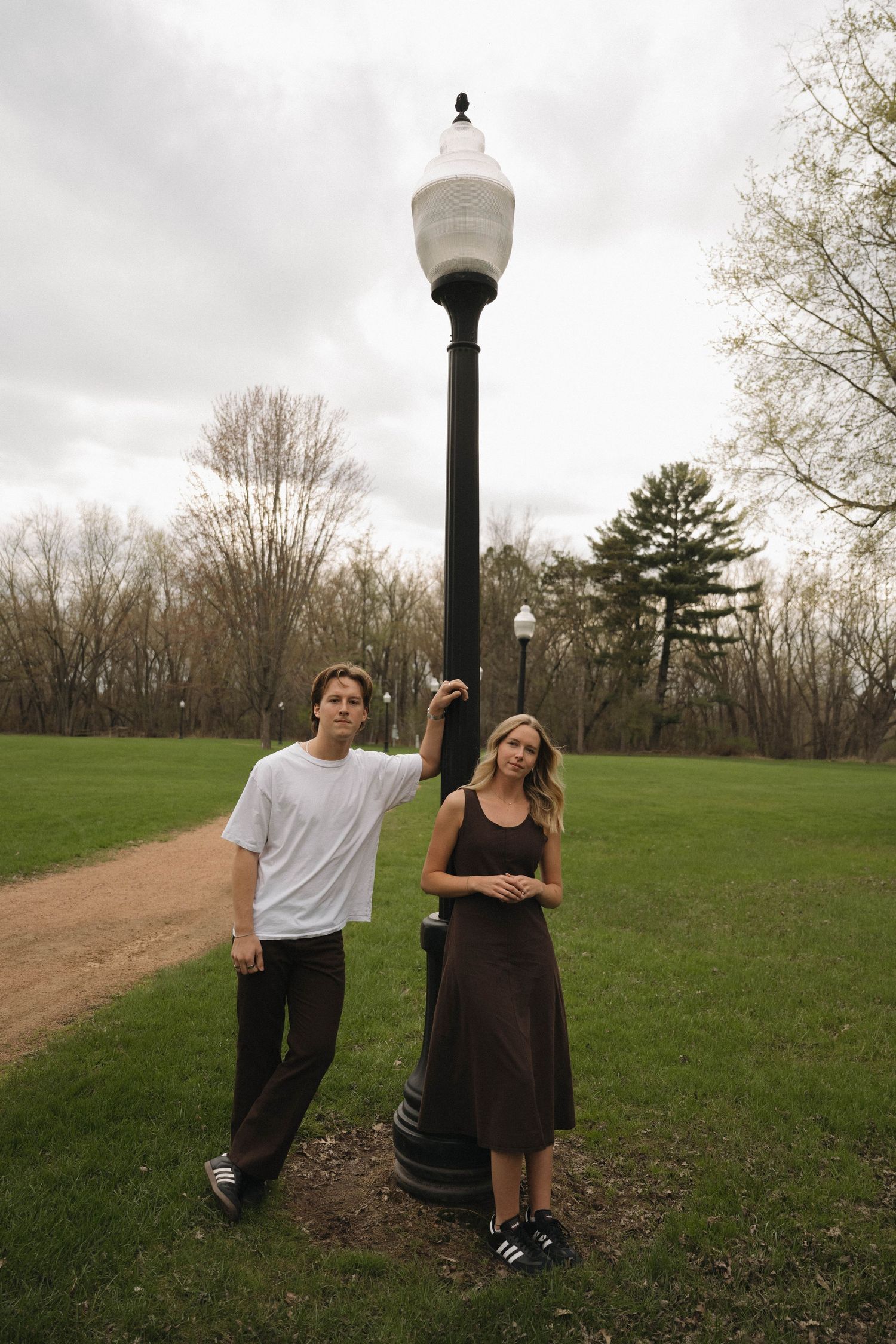 A man and woman standing by a black lamp post in a park, wearing casual outfits, with green grass and trees behind.