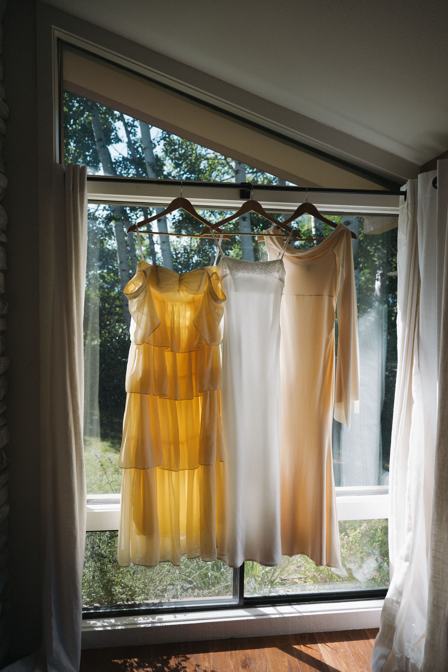 Three wedding dresses hanging on wooden hangers in sunlit window, including yellow, white, and champagne gowns.