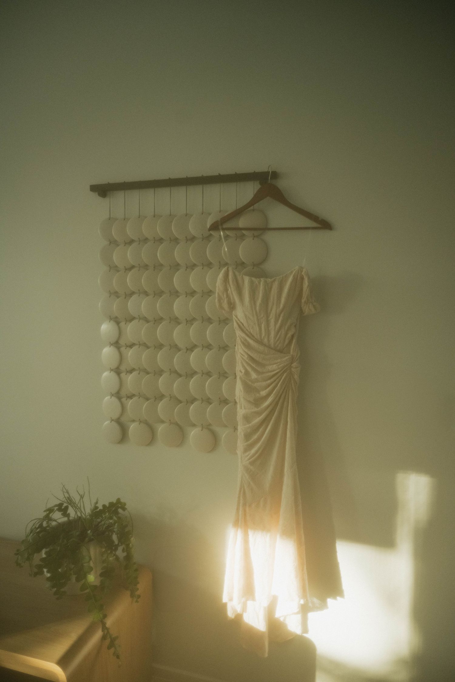 White wedding dress hanging on a wooden hanger against a wall decorated with circular ornaments in soft light.