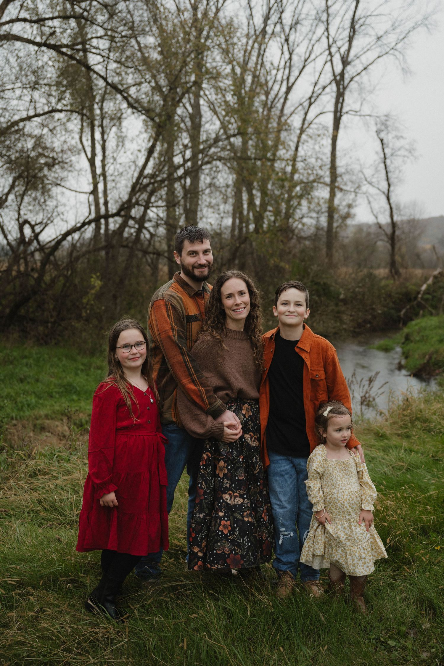 A family of five posing together near a creek surrounded by bare trees in autumn.