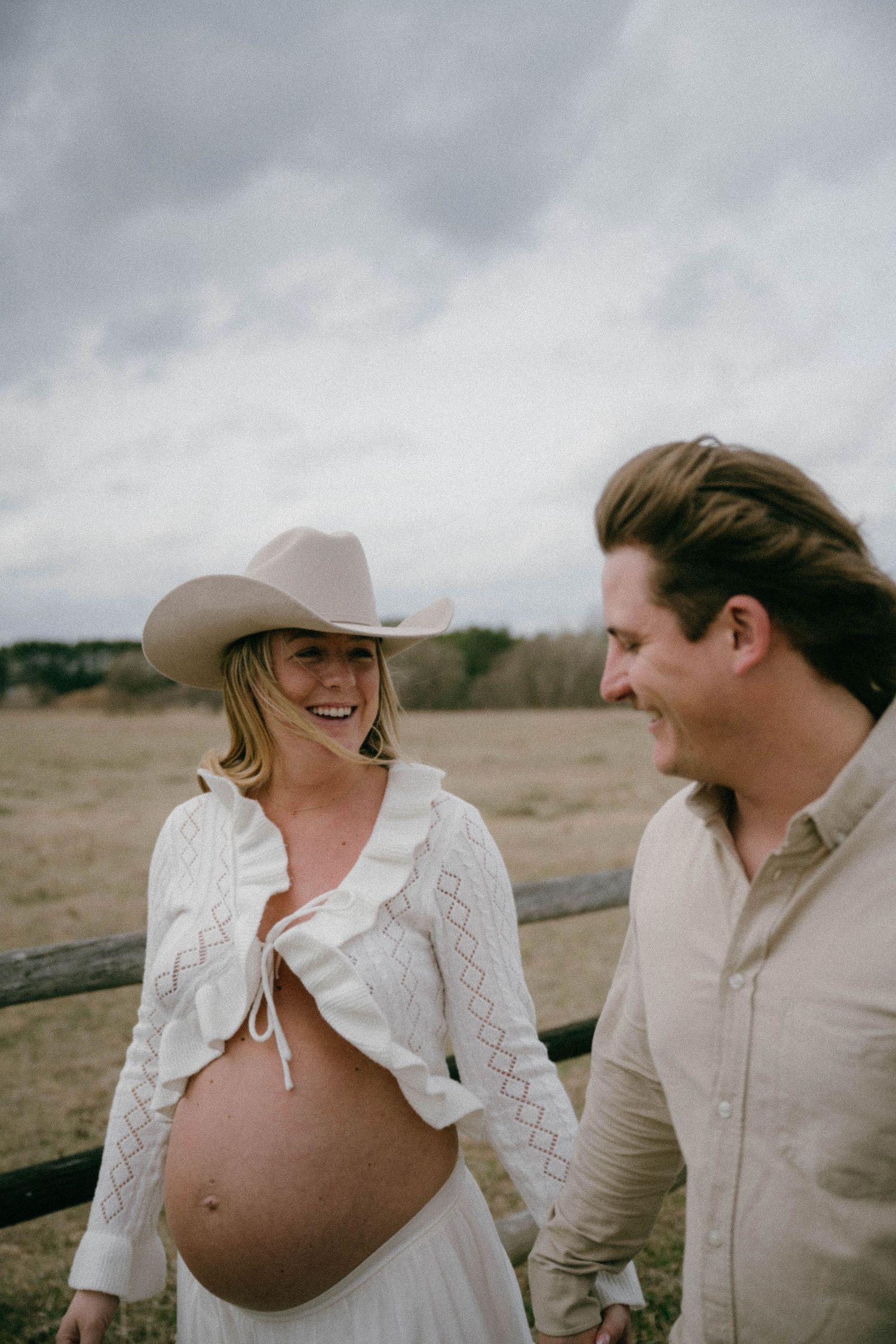 Laughing pregnant woman in cowboy hat and white crop top with a man in an open field on cloudy day.