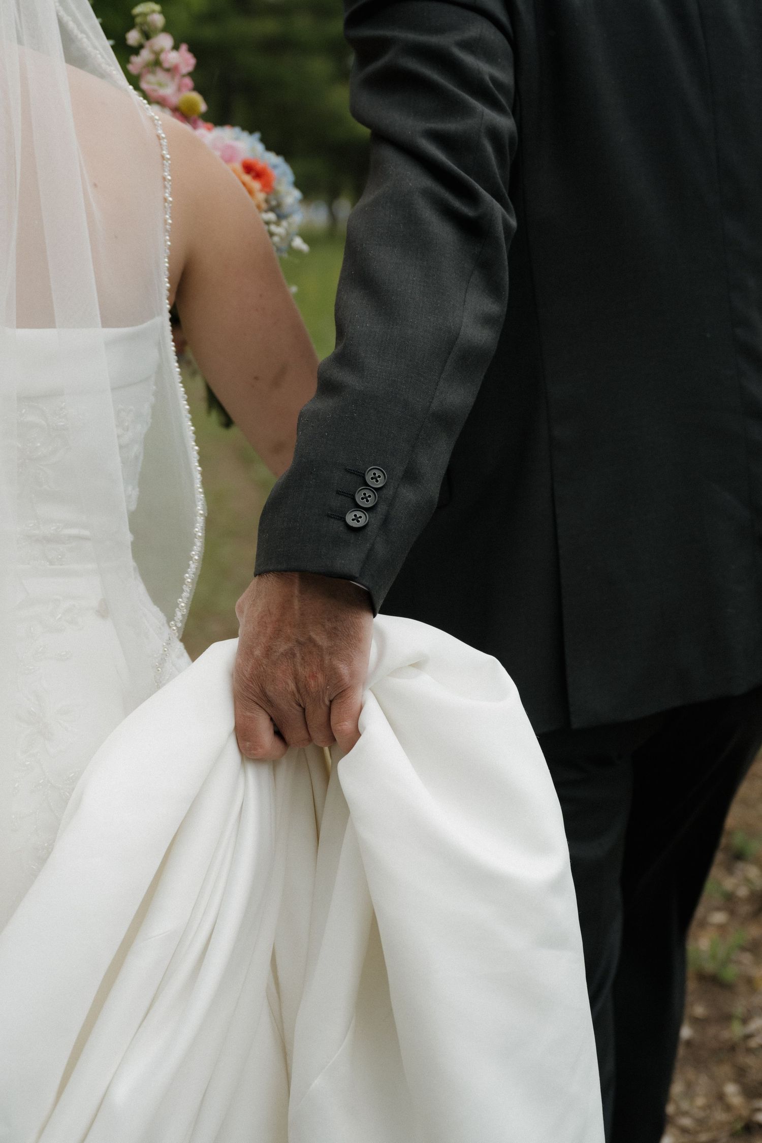 A groom in a black suit gently holds the train of a bride's white wedding gown as they walk together outdoors.