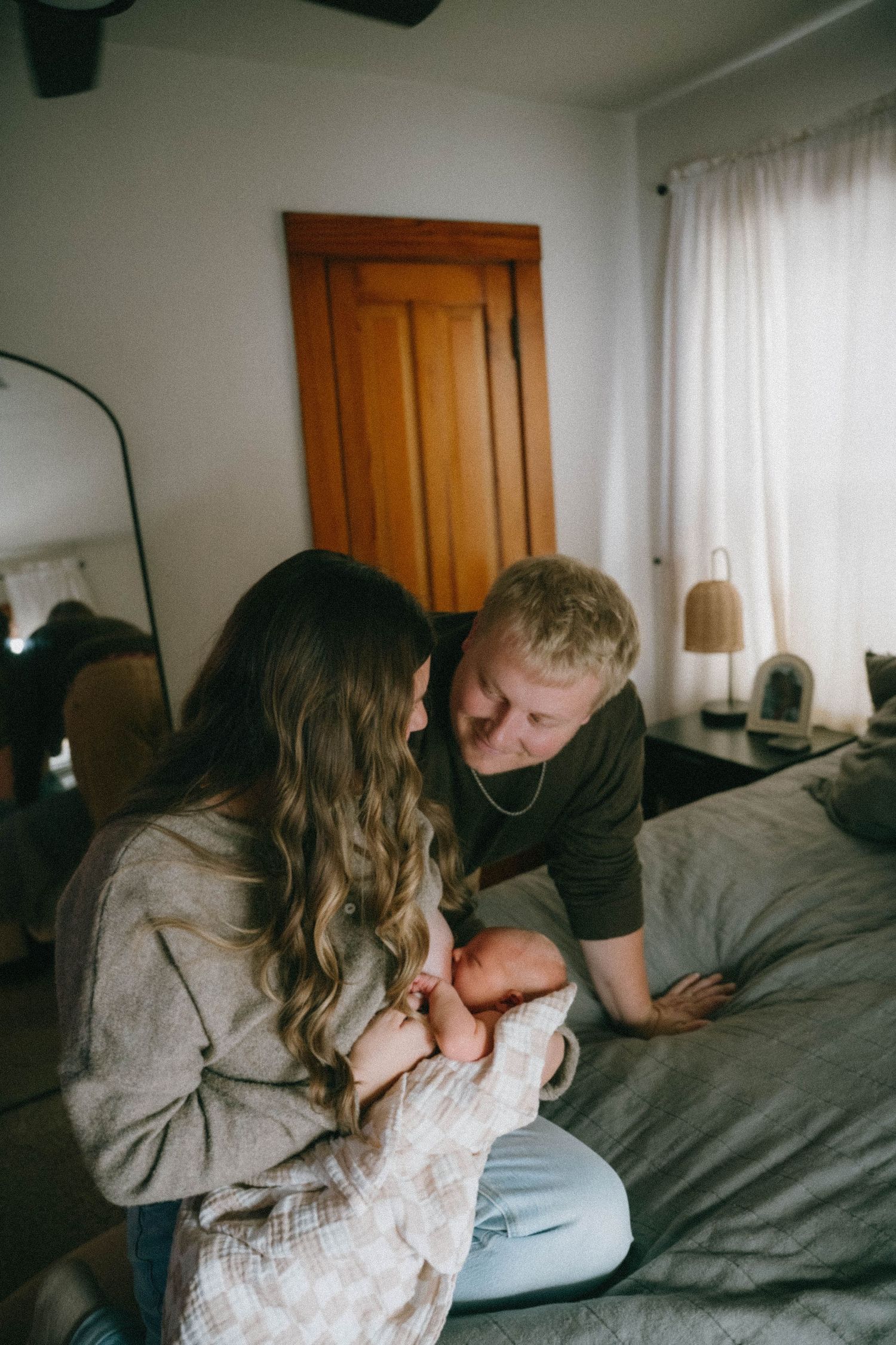 New parents sitting on bed gazing lovingly at their newborn baby in cozy bedroom.
