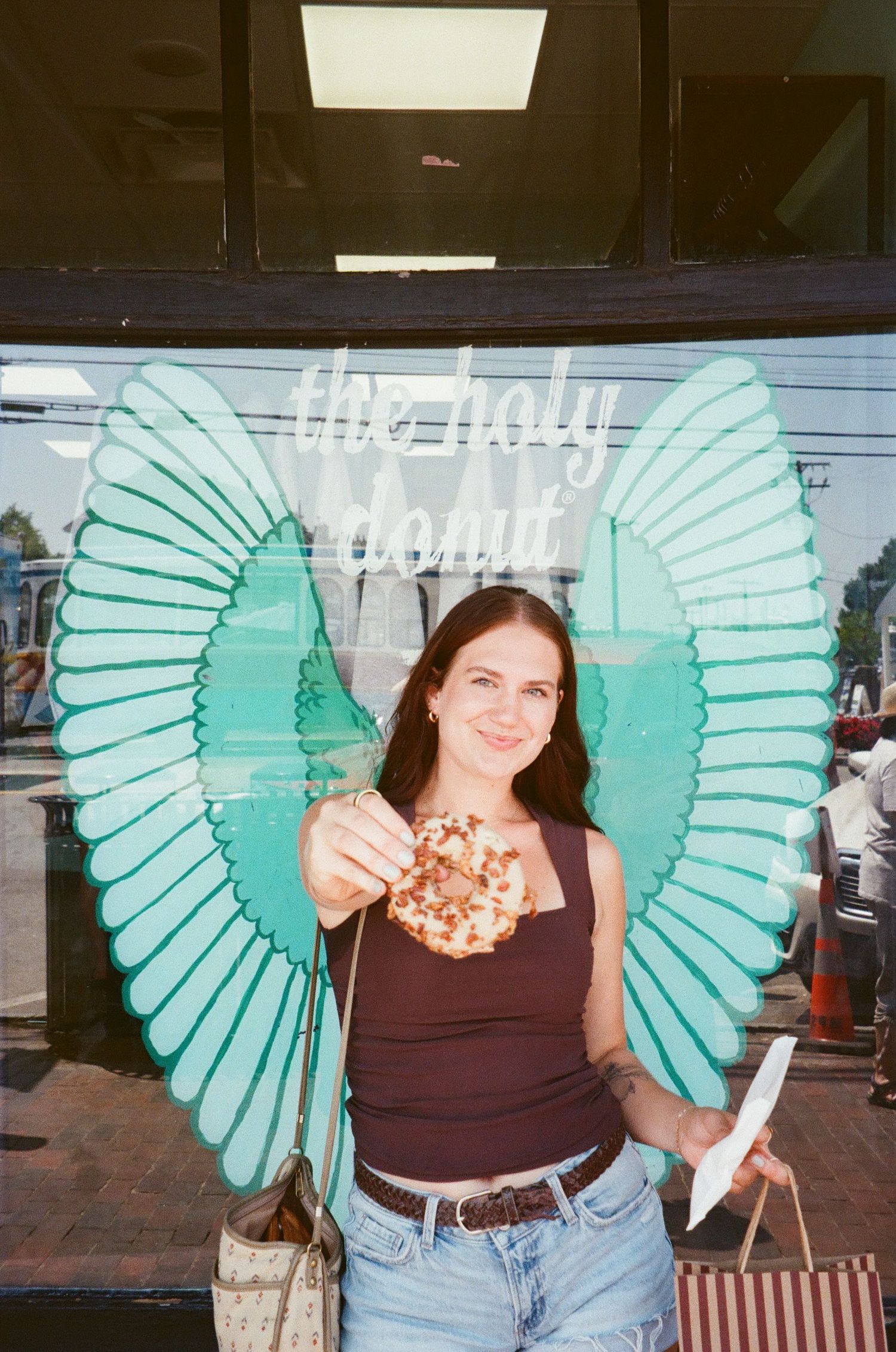 Brunette woman holding donut in front of teal angel wings mural at The Holy Donut shop.