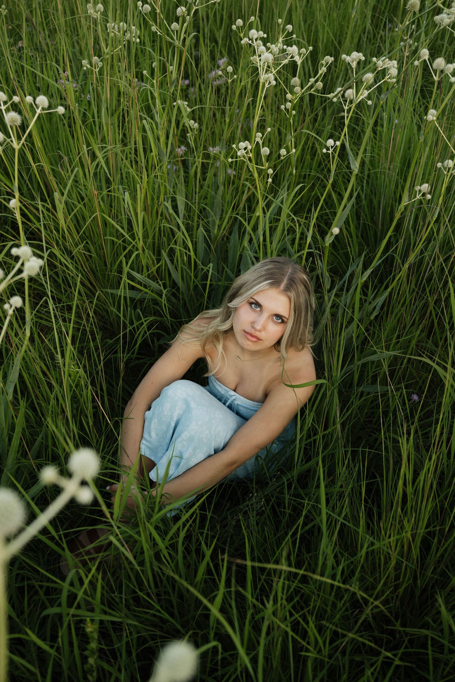 A blonde woman in a light blue dress sits surrounded by tall green grass and white wildflowers outdoors.