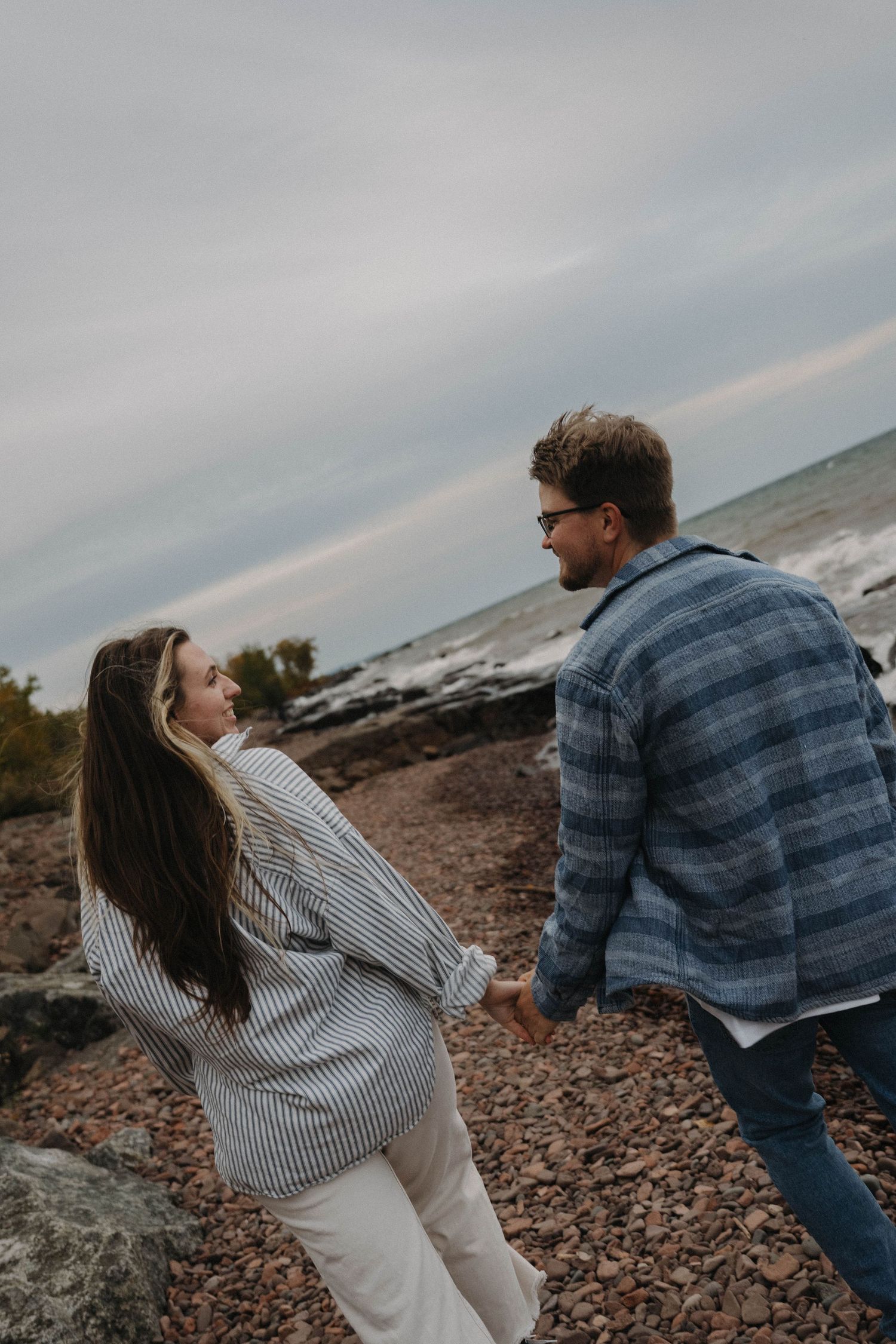 Couple holding hands walking along rocky shoreline with overcast sky.