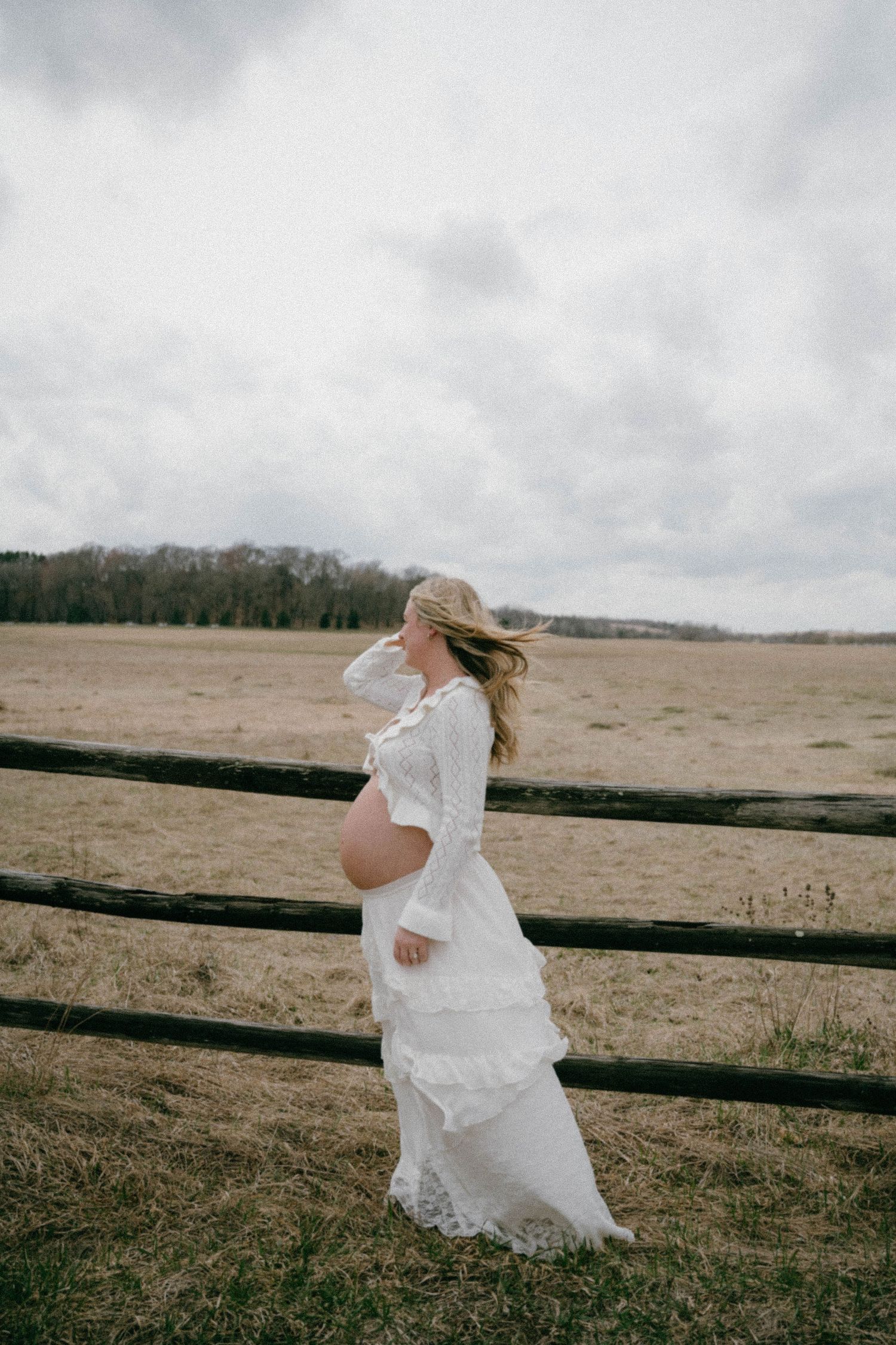 Pregnant woman in a white lace dress standing by a wooden fence in an open field on a cloudy day.