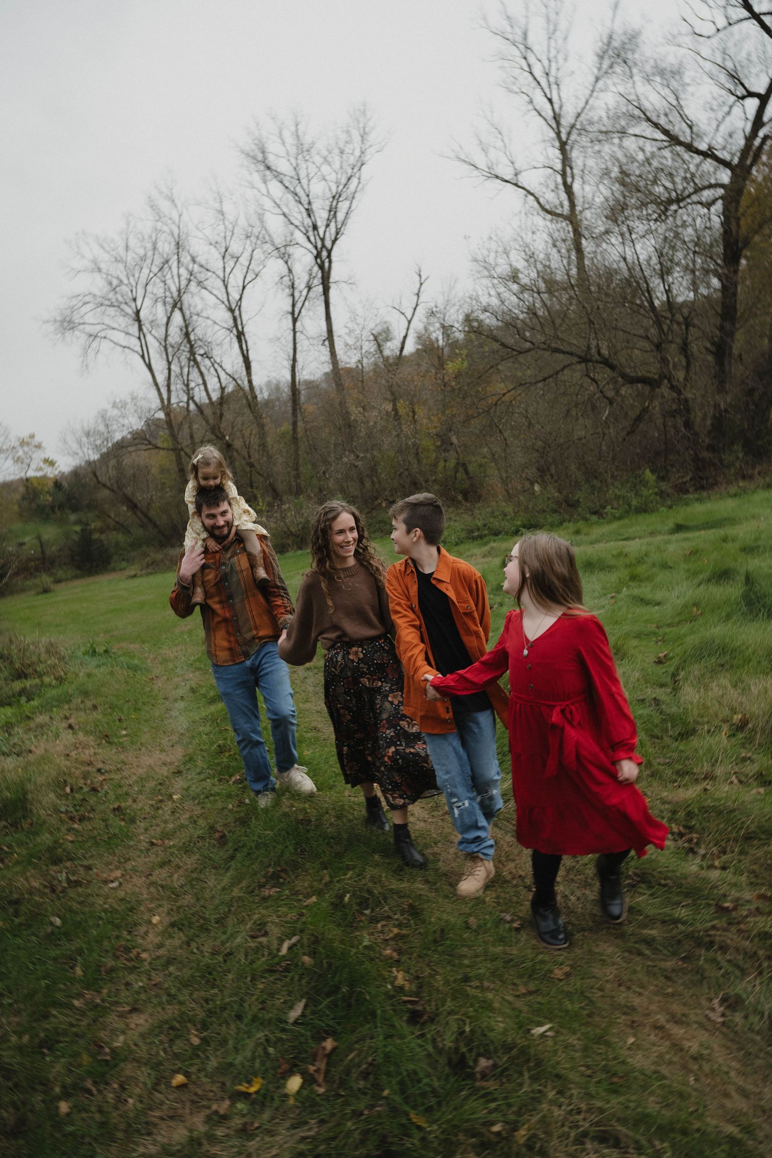 Family of five walks together along a grassy path through an autumn landscape with bare trees in background.