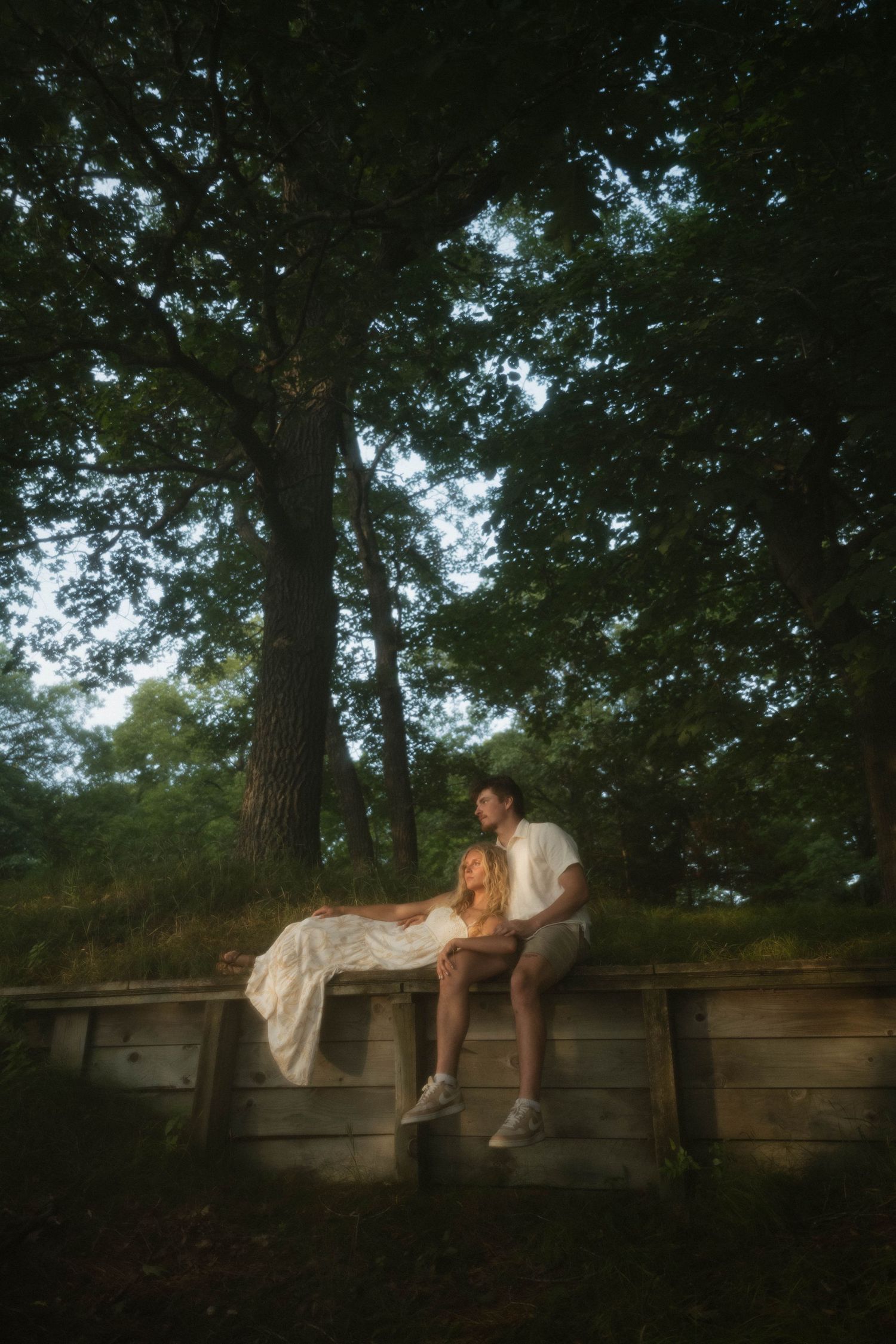 Couple sitting on rustic wooden bench under large oak tree in lush green park setting.