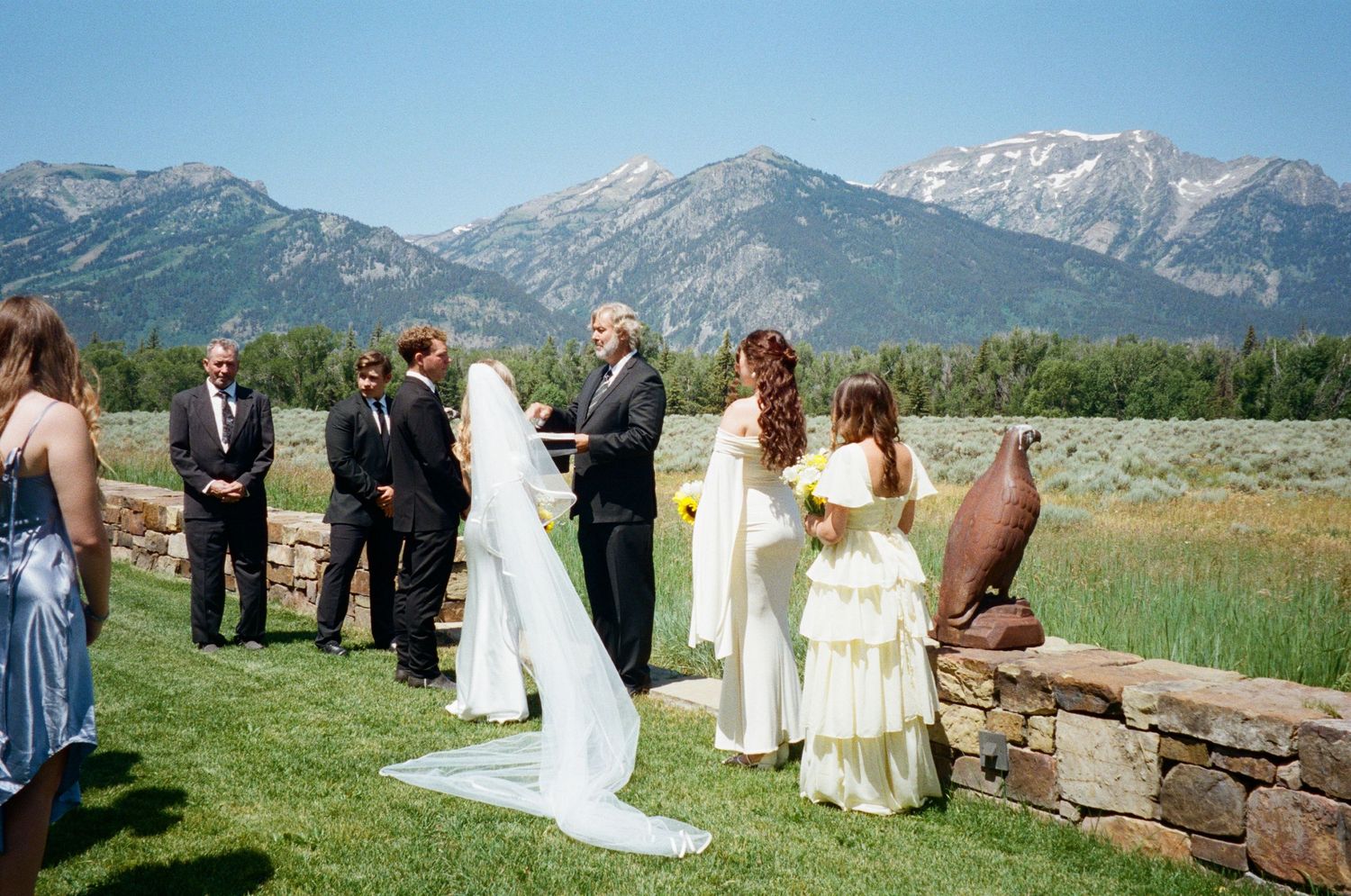 Outdoor wedding ceremony with bride in flowing veil before stunning snow-capped mountain landscape.