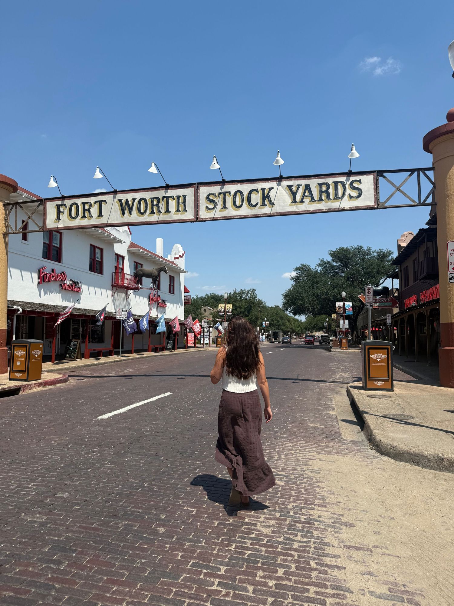 Woman walking under Fort Worth Stock Yards sign on sunny brick street in historic Texas district.