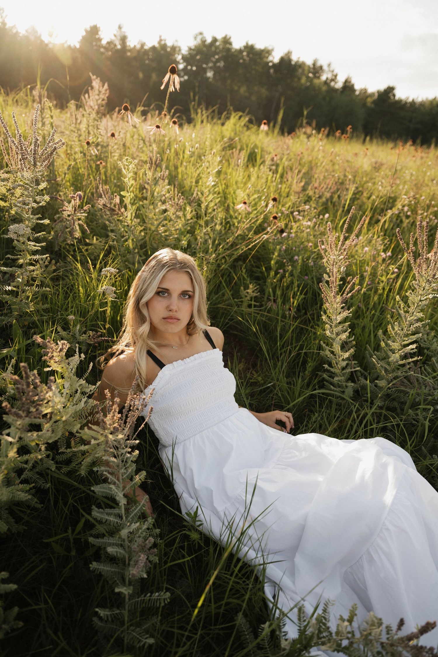 A blonde woman in a white dress reclines gracefully among tall wildflowers in a sunlit meadow at sunset.