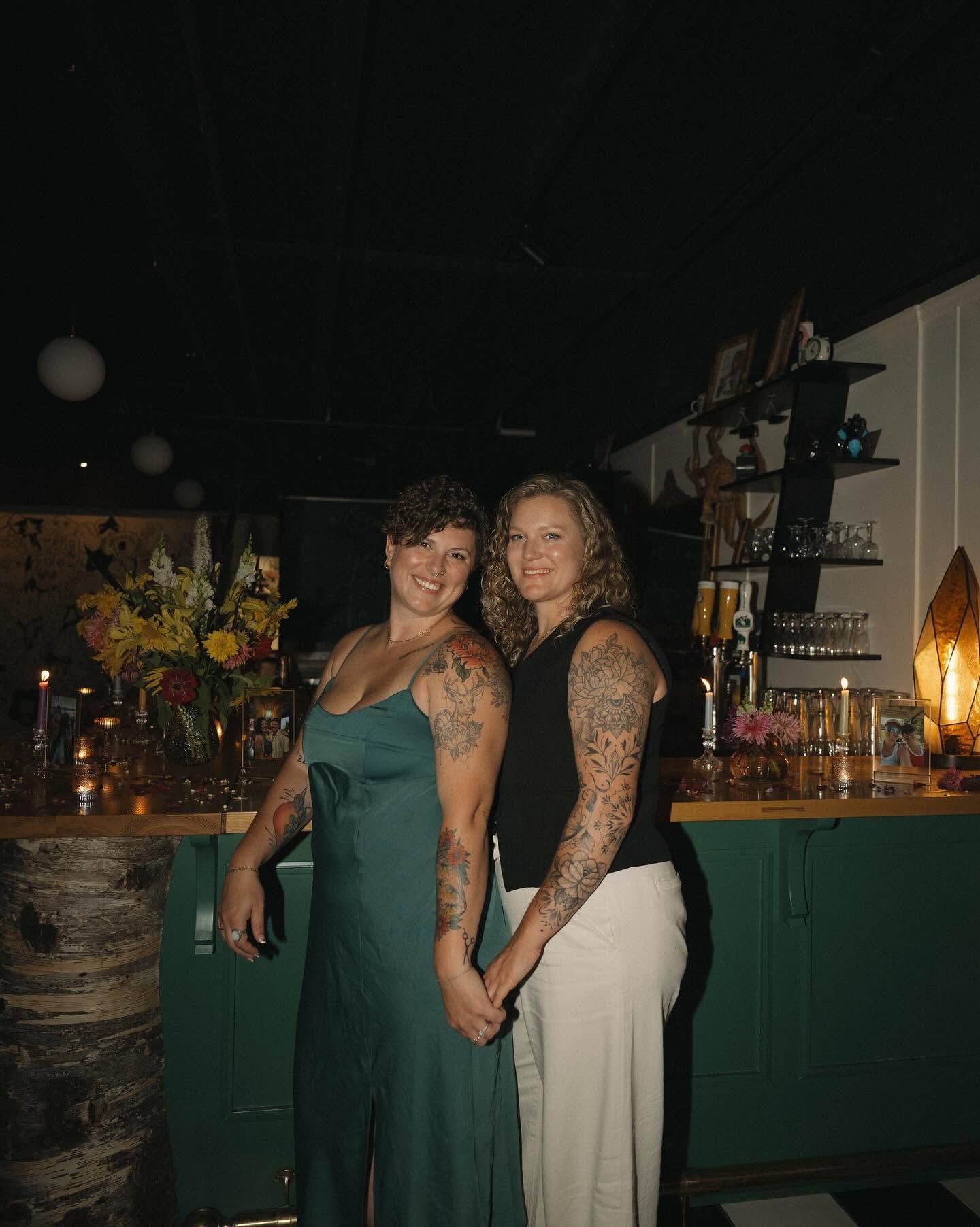 Two tattooed women posing together in a bar with green cabinetry, one in a green dress, one in black top and white pants.
