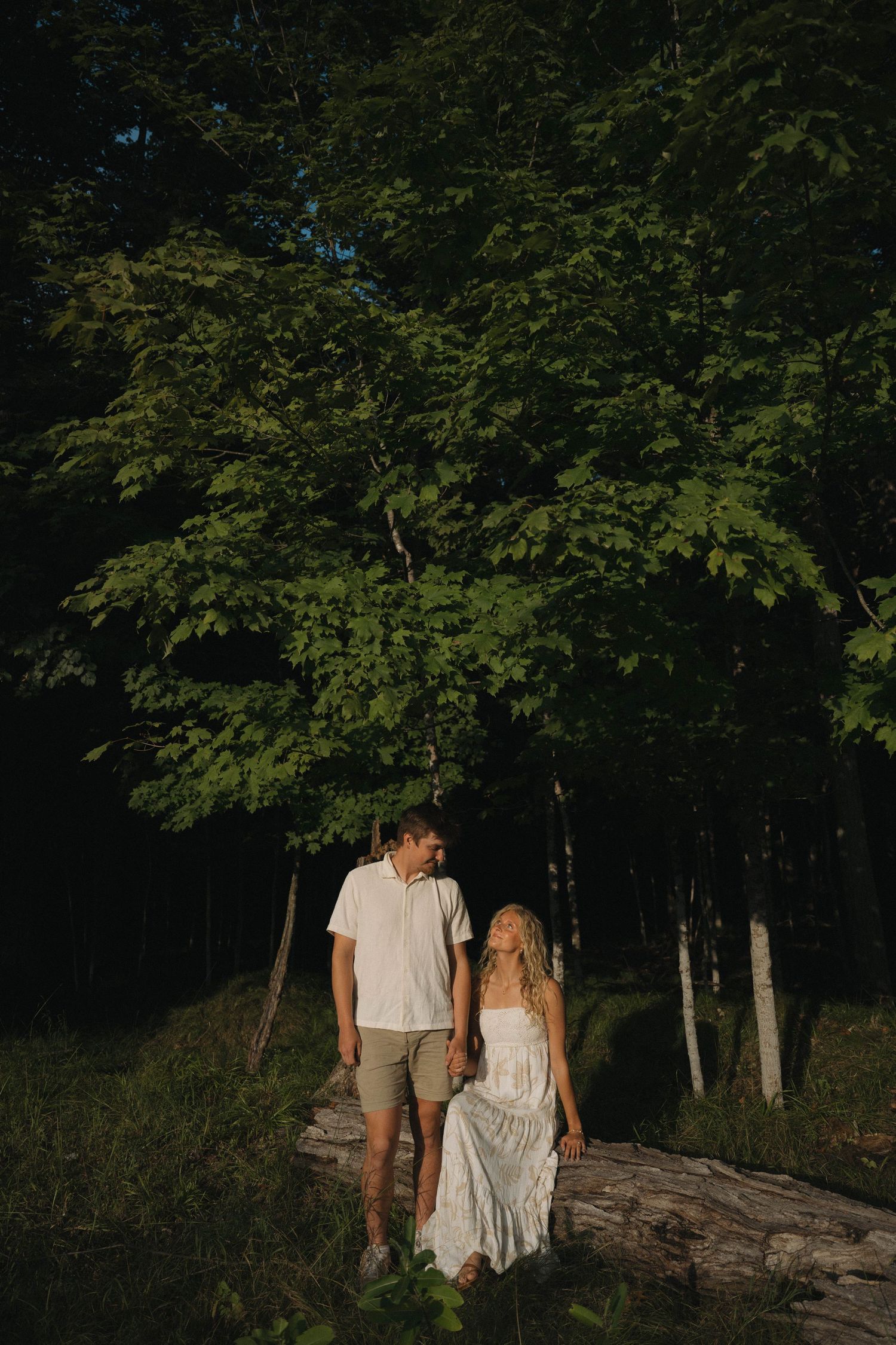 Couple in white outfits holding hands in a sunlit forest clearing, standing on a fallen log surrounded by lush green trees.