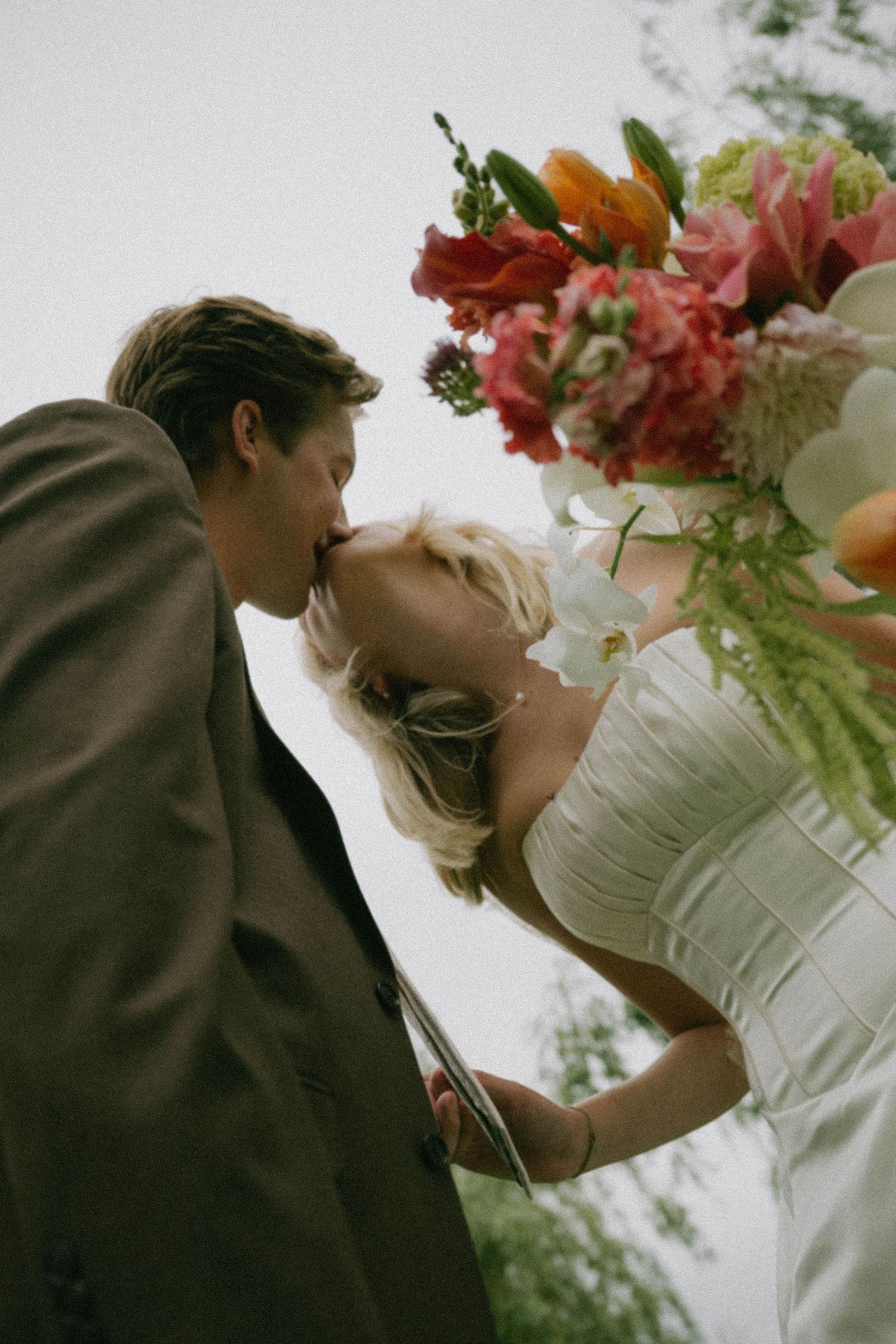 Bride and groom sharing a romantic kiss beneath a vibrant colorful wedding bouquet of pink and orange flowers.
