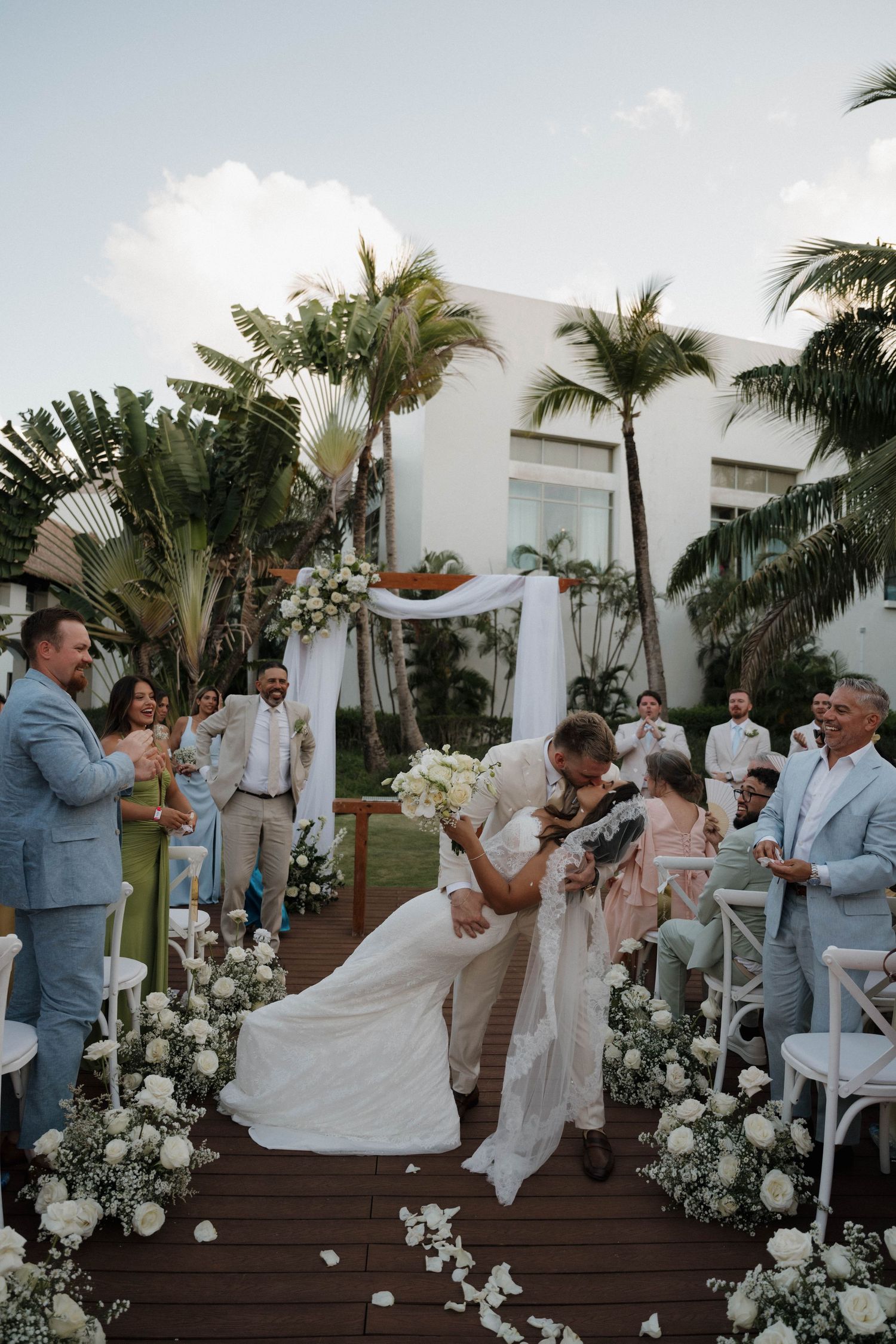 Bride and groom sharing romantic dip kiss at outdoor tropical wedding ceremony surrounded by white floral arrangements.