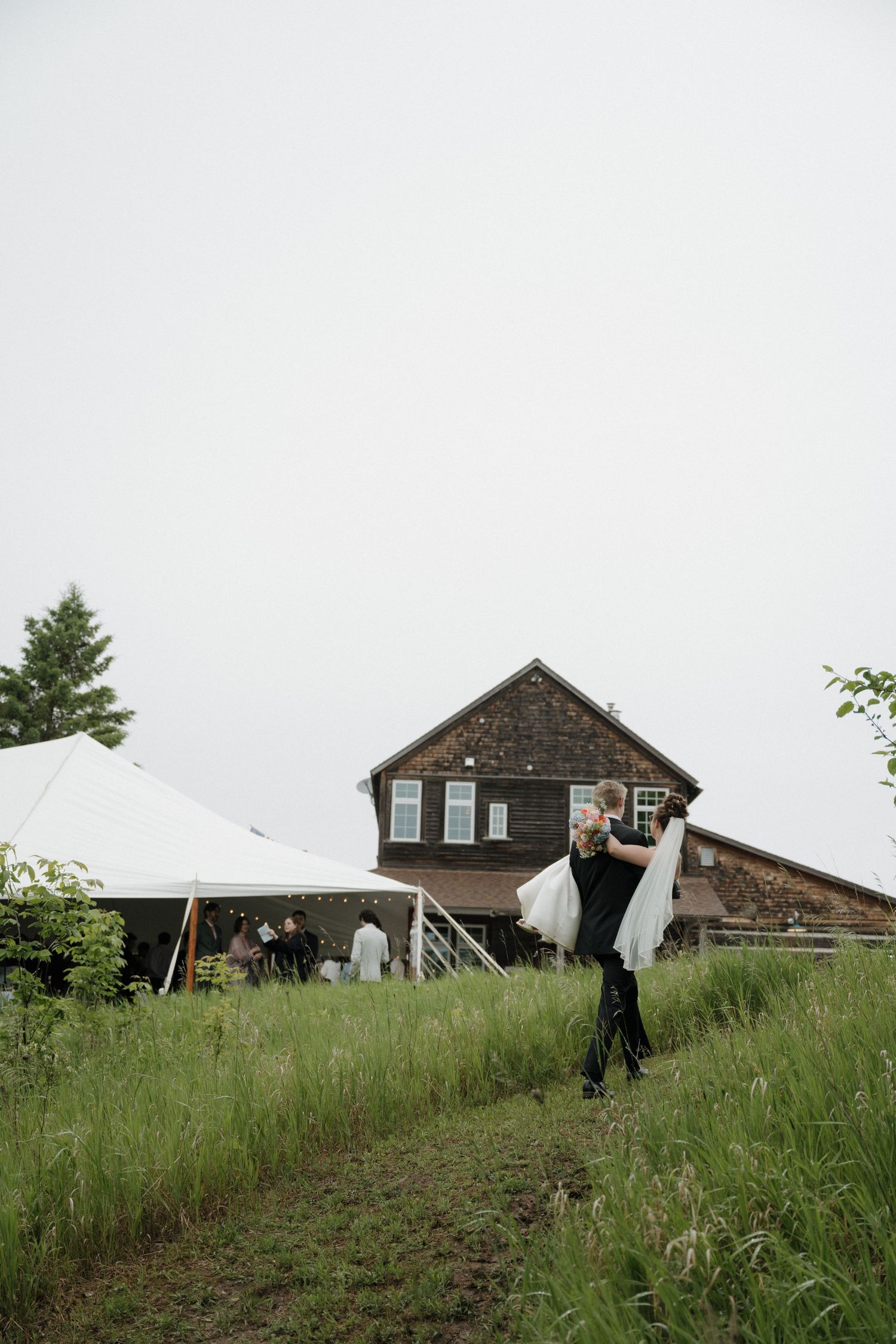 Groom carries bride in white dress near rustic barn wedding venue.