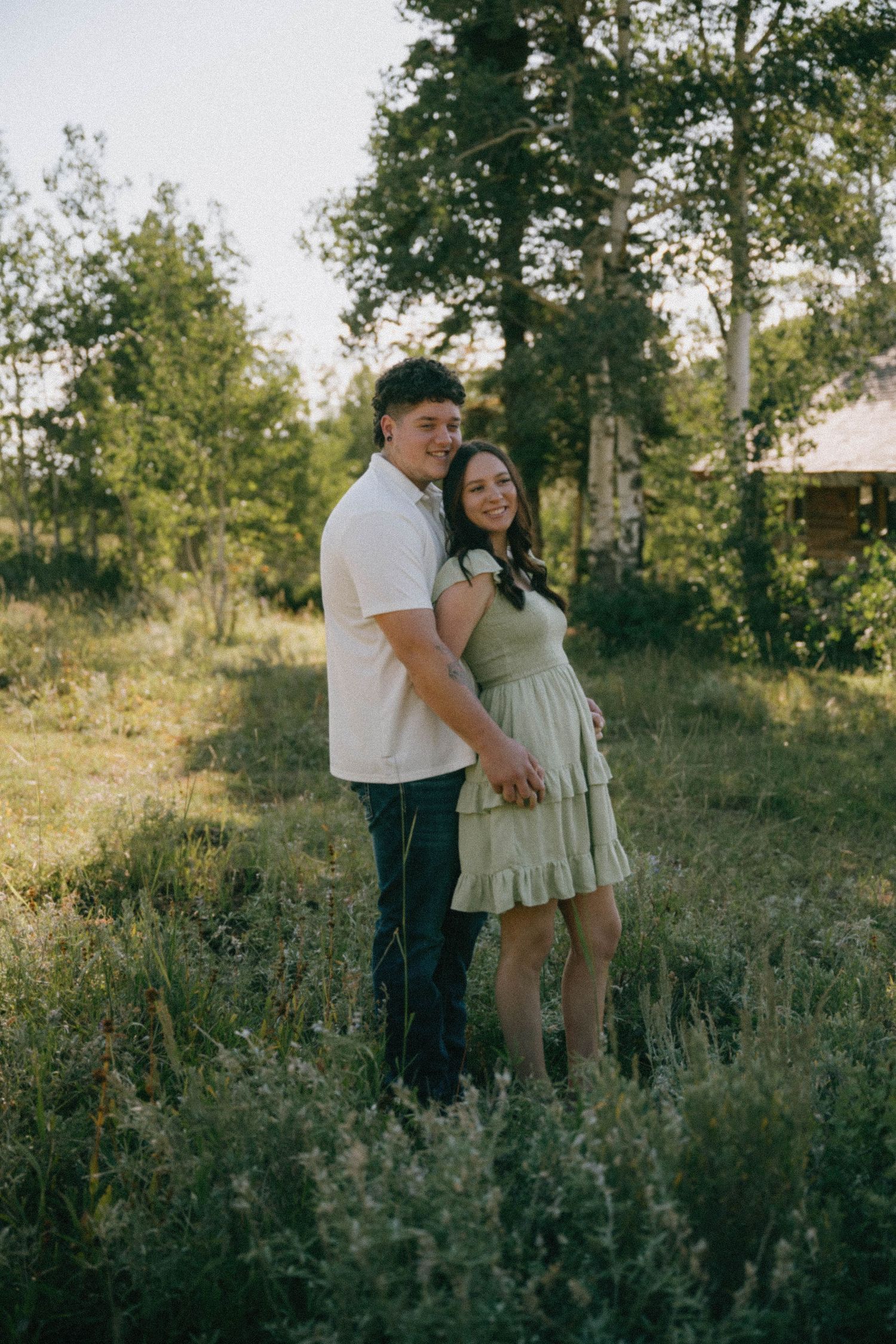 A smiling couple holds hands while standing in a wildflower meadow surrounded by green trees at golden hour.