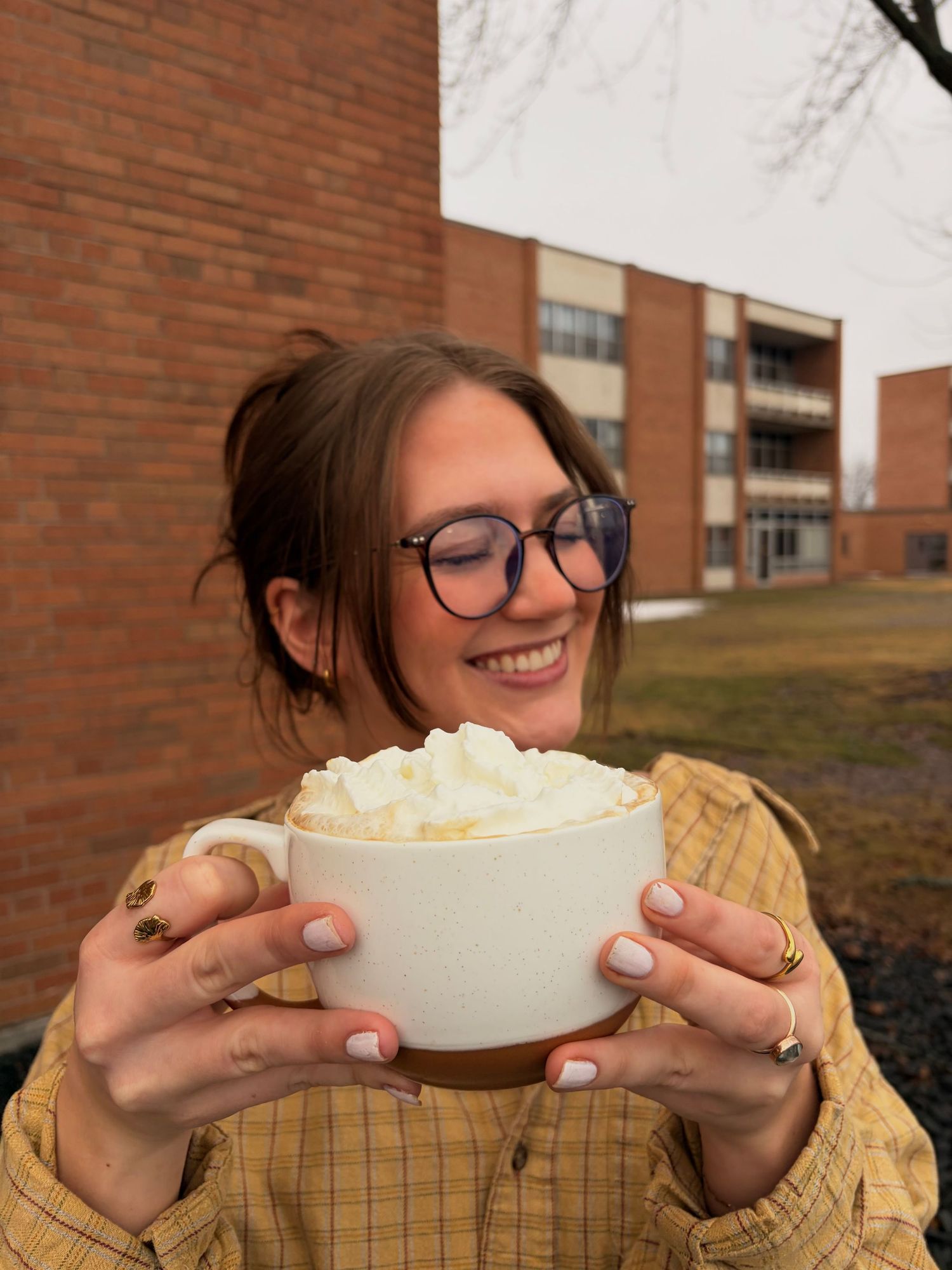 Smiling woman with glasses holding large white mug topped with whipped cream outdoors.