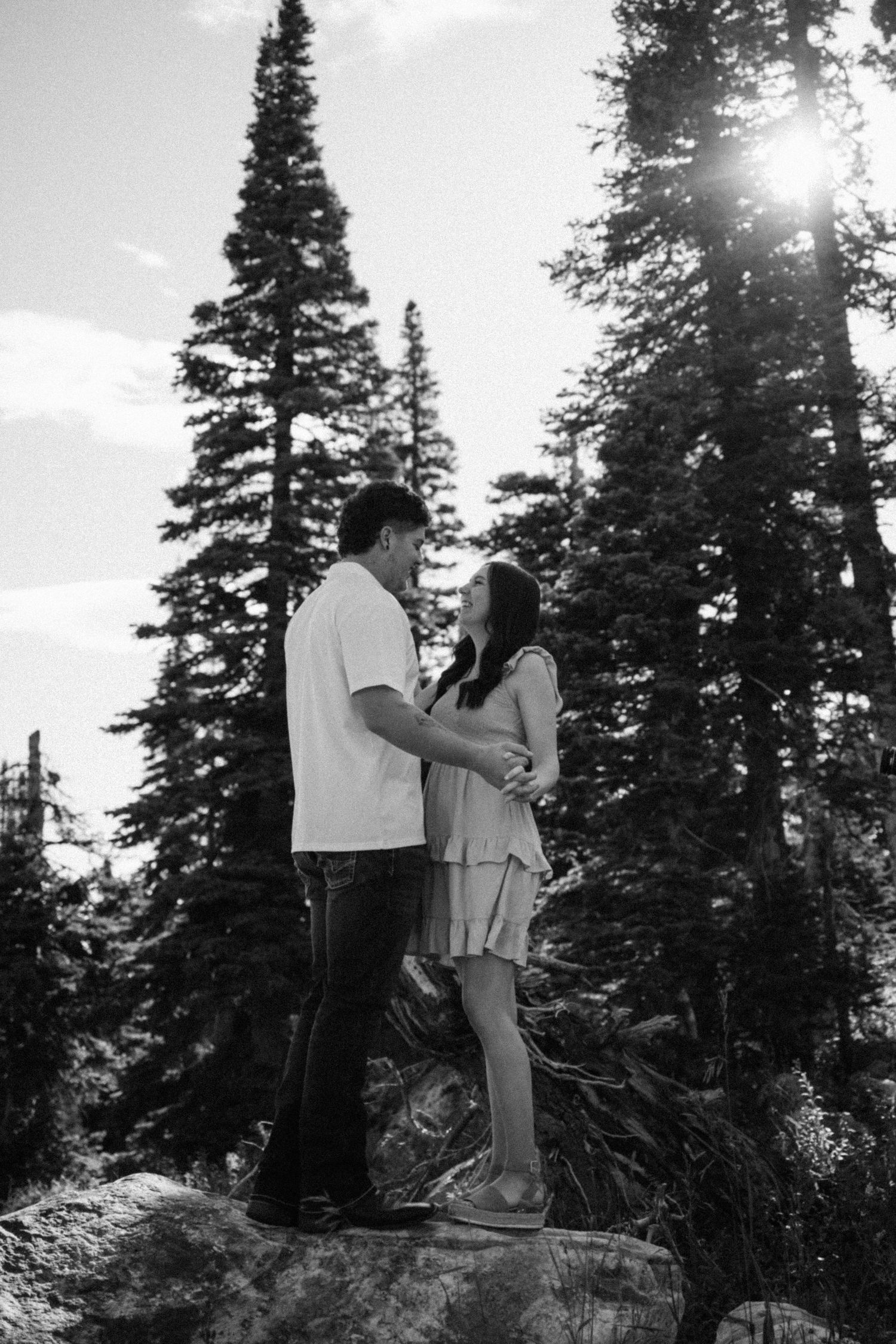 Black and white photo of a couple standing on rocks surrounded by tall pine trees in a mountain forest.