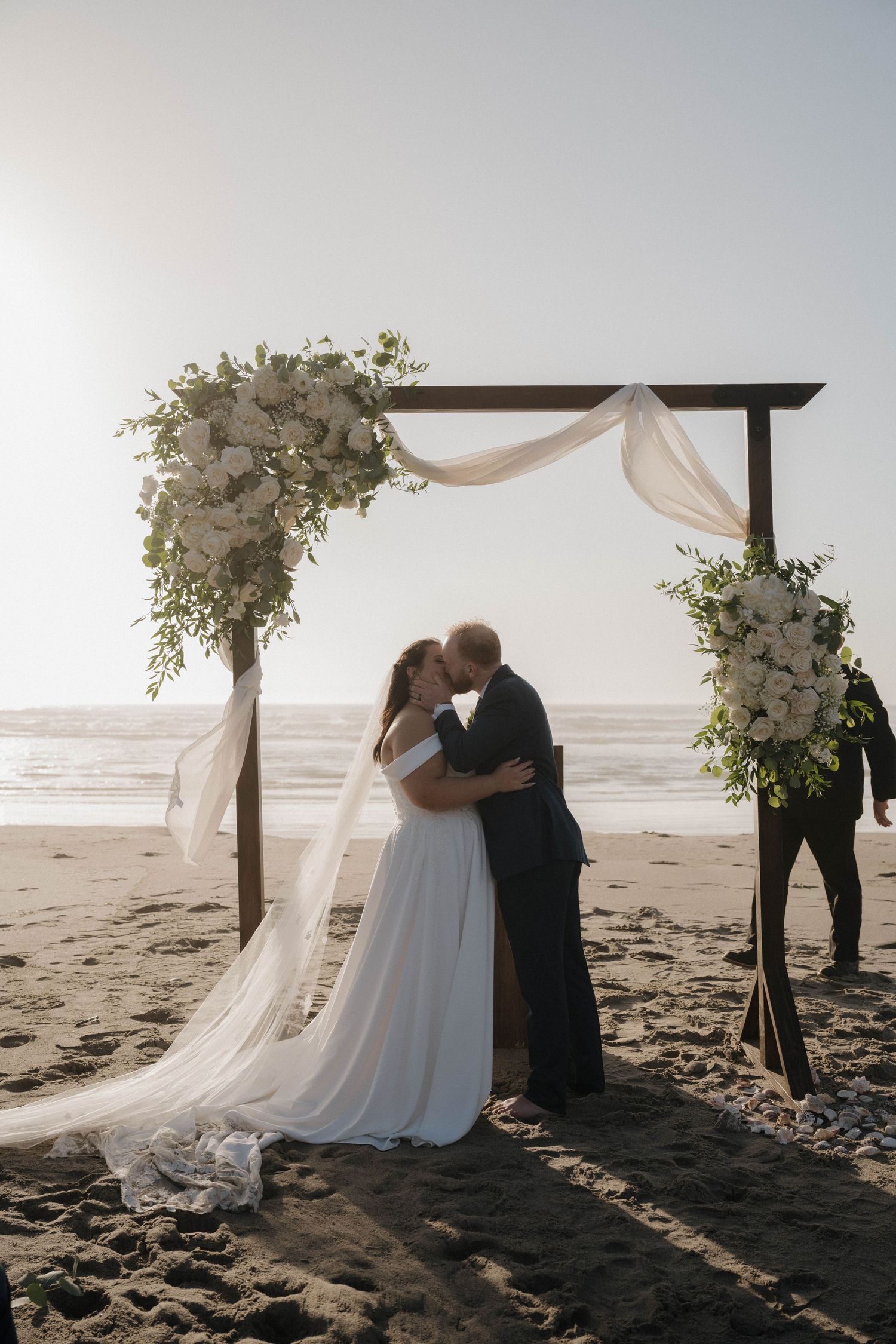 A bride and groom share their first kiss under a floral wooden arch at a beach wedding.