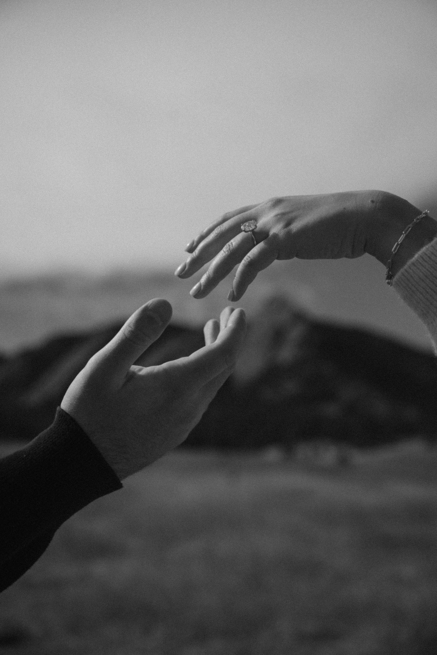 Black and white close-up of two hands reaching toward each other with engagement ring visible.