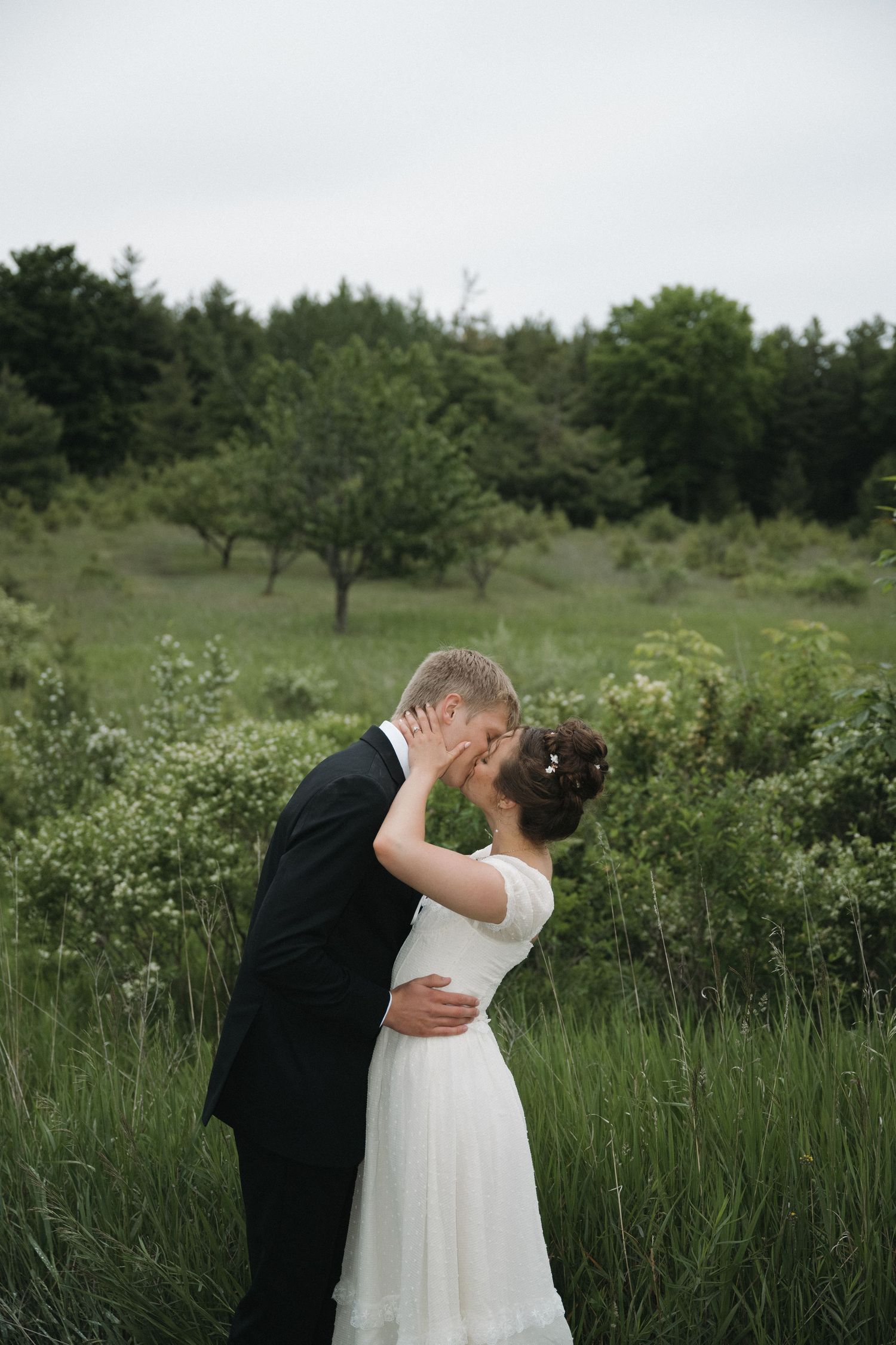 A bride and groom share a romantic kiss in a lush green meadow, with the bride in a white dress and groom in a black suit.