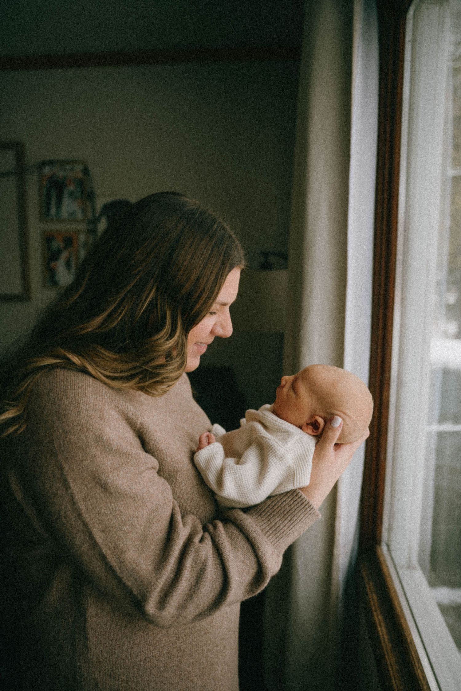 A mother gazes lovingly at her newborn baby while standing beside a window in soft natural light.