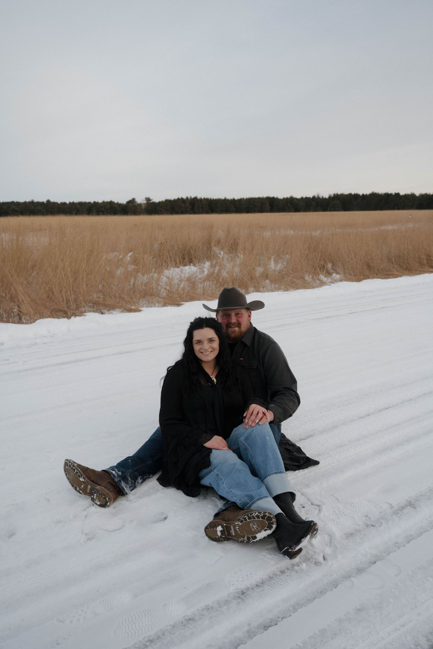 Couple sitting together on snowy road in winter landscape, man in cowboy hat, woman in black coat.
