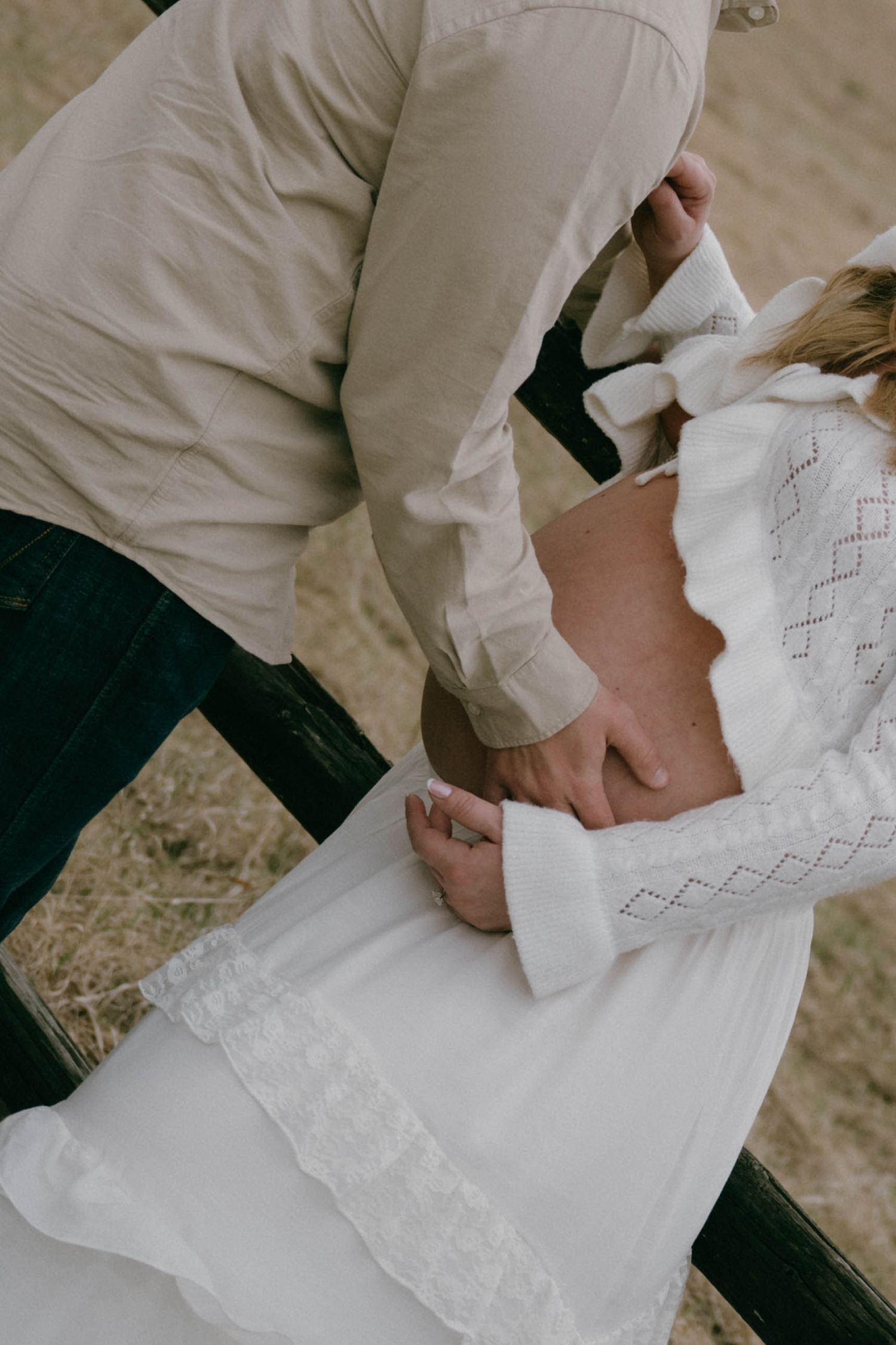 A couple sharing an intimate moment, the woman in a white lace dress holding hands with a man in a beige shirt outdoors.