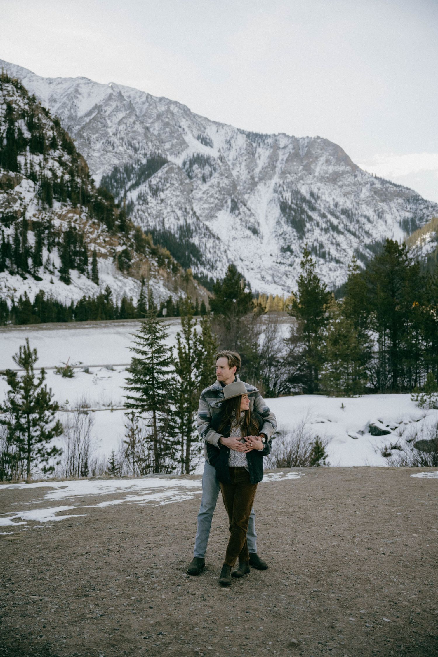 Couple embracing in a snowy mountain landscape with pine trees and a frozen lake in the background.