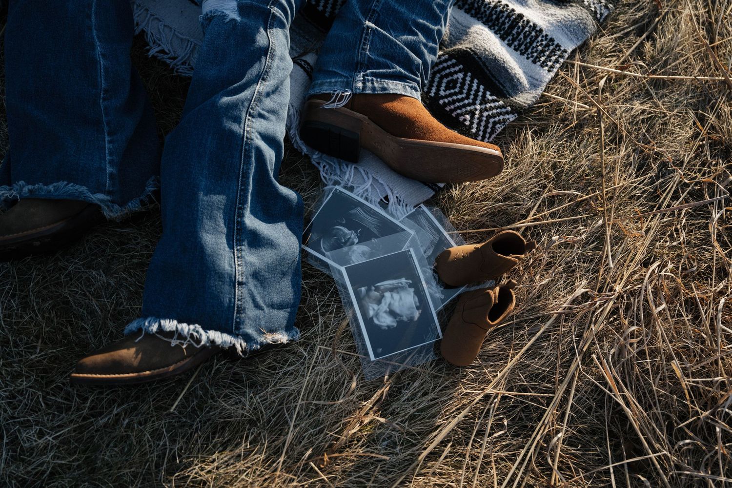 Couple's feet in jeans and brown shoes on dry grass with ultrasound photos between them.