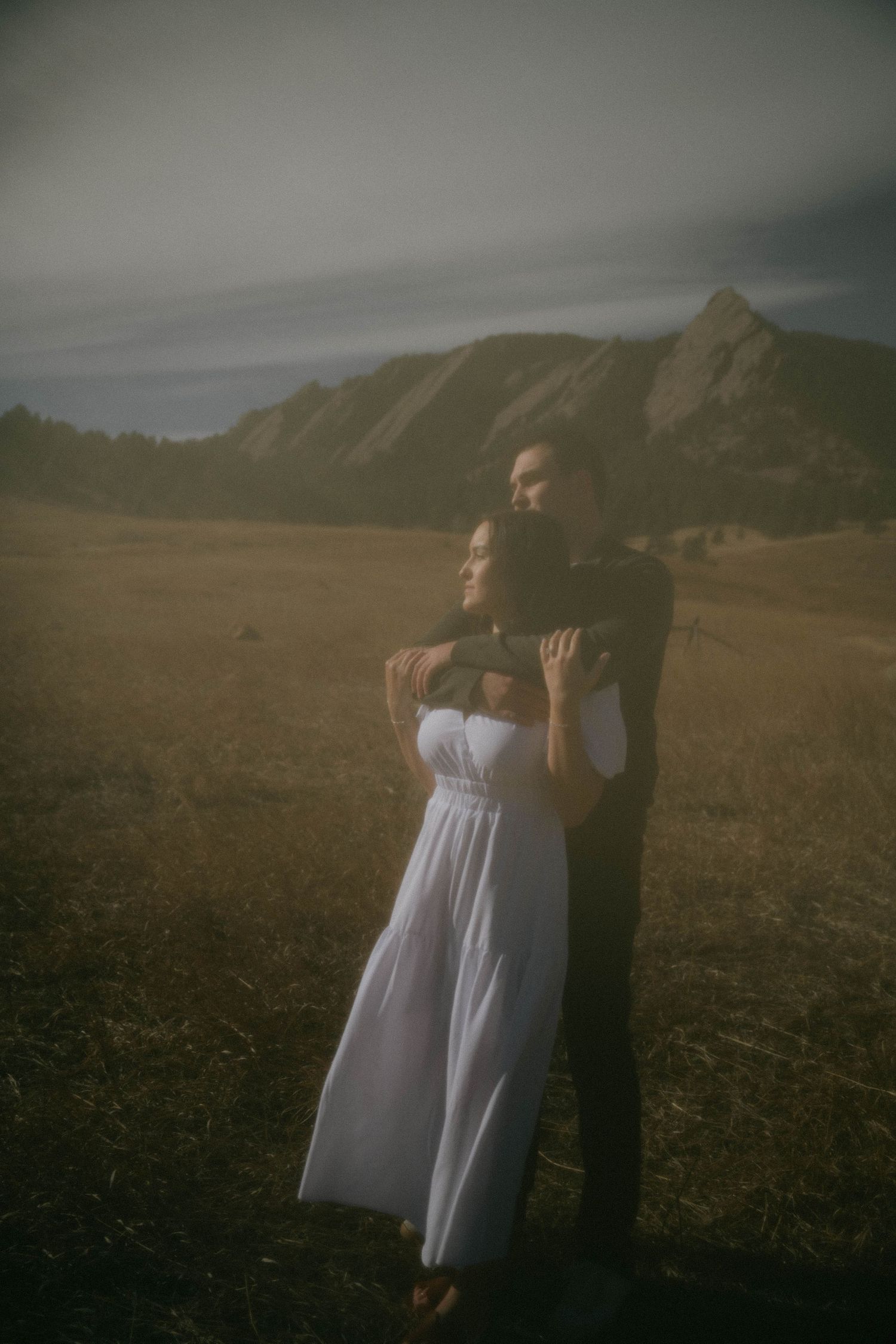 Couple embraces in golden meadow at dusk with dramatic Flatiron mountains in background.