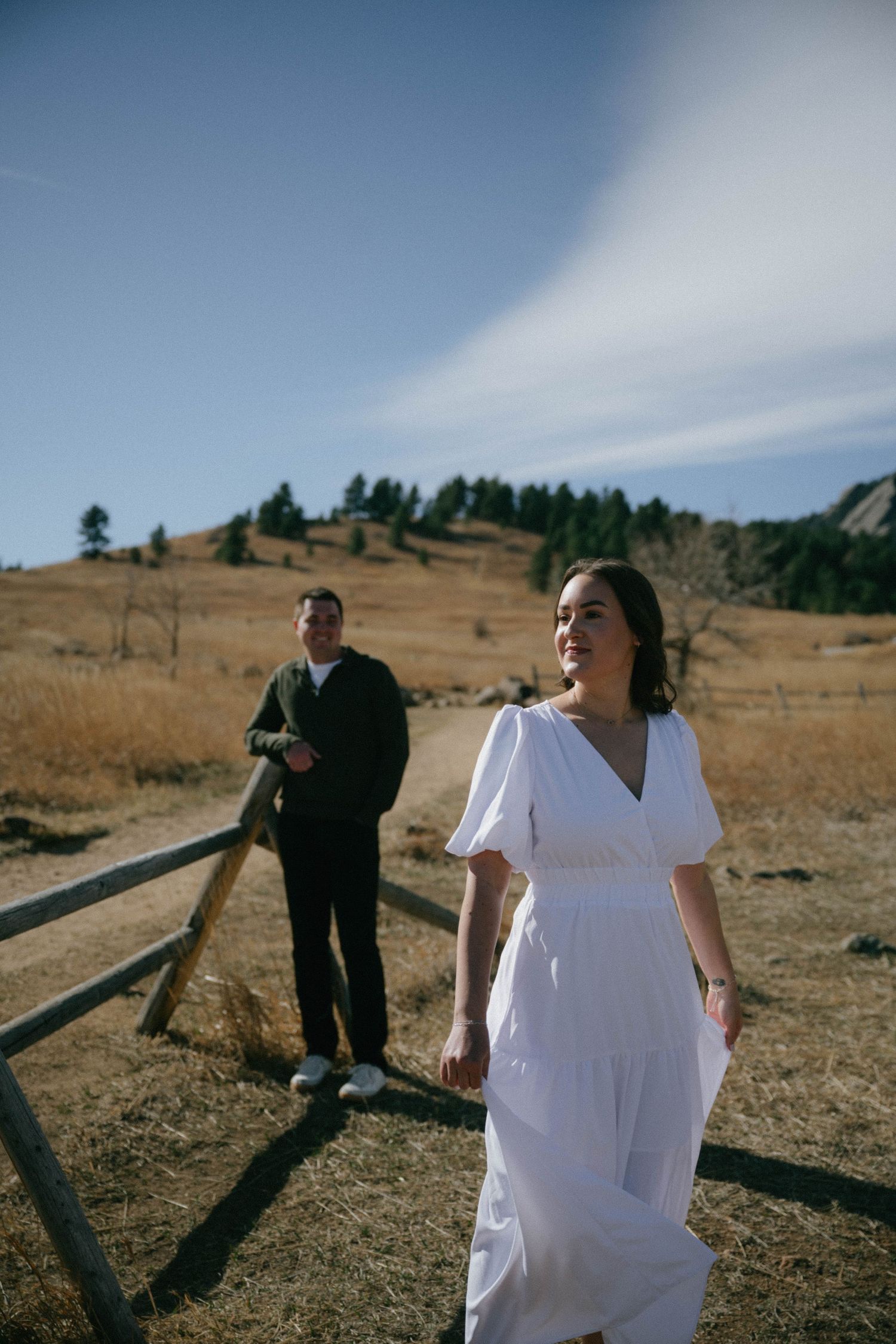 Woman in white dress walks path at Chautauqua Park with man and Flatirons mountains in background.