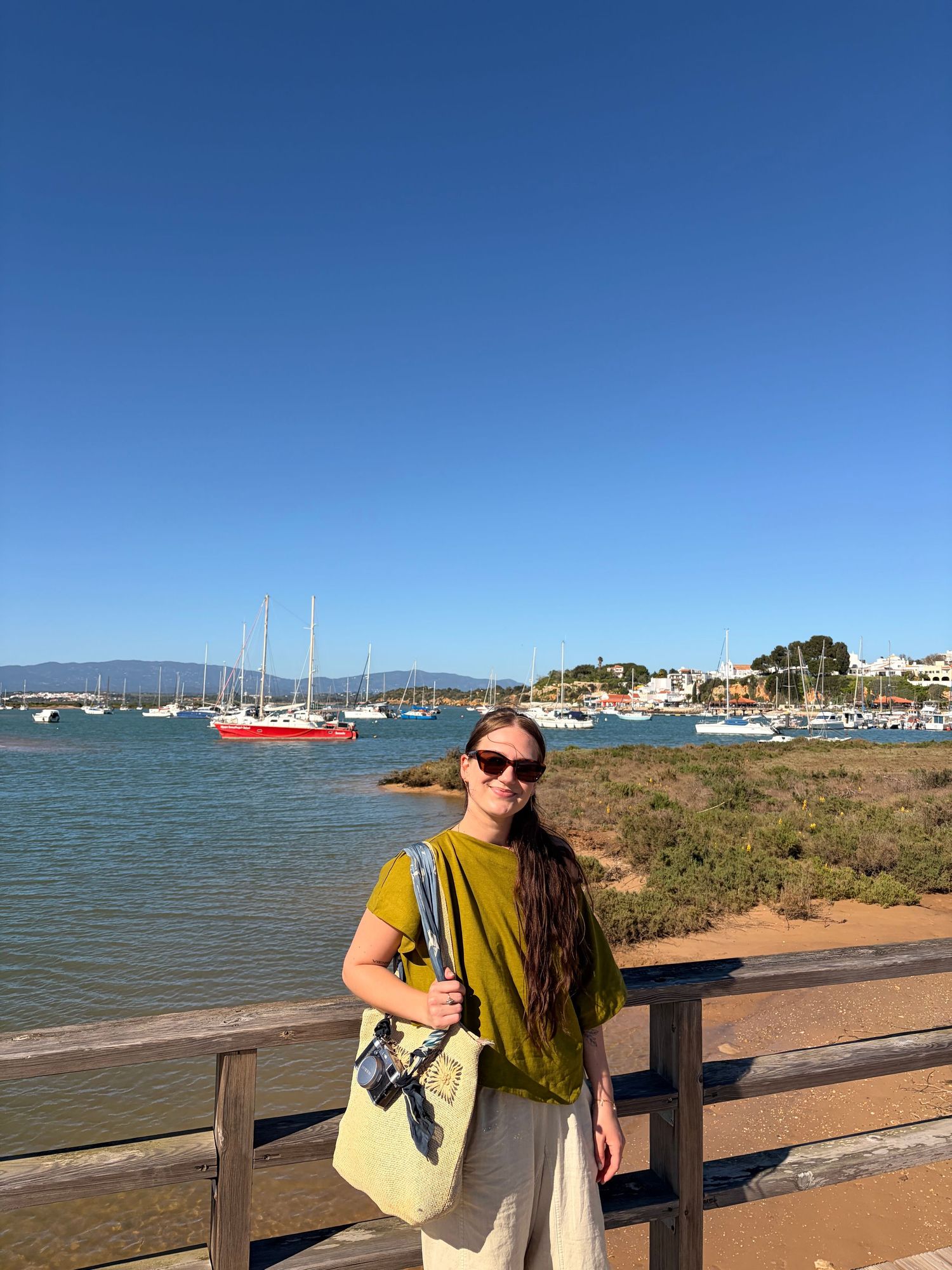 Woman in olive top with tote bag stands on waterfront boardwalk with sailboats and blue sky behind her.