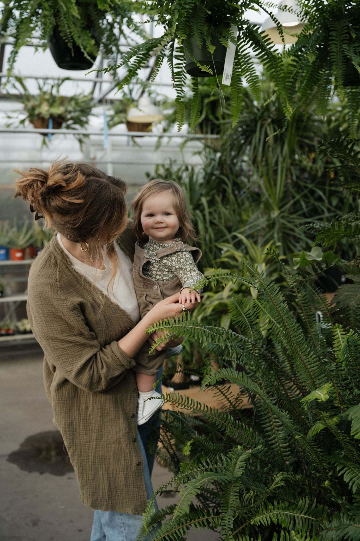 Mother holding toddler girl while exploring lush green plants in a greenhouse nursery.