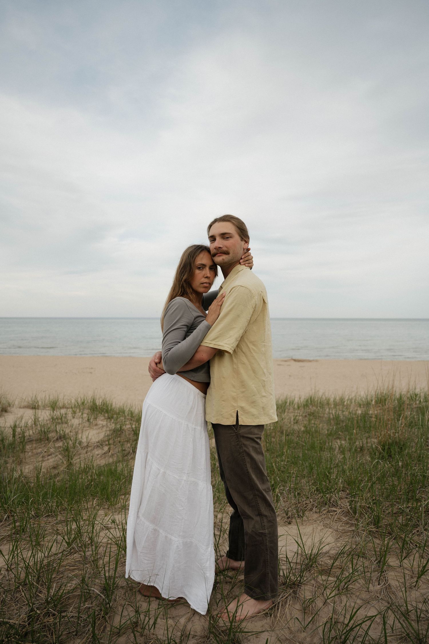 Couple embracing on a sandy beach with ocean backdrop, woman in white skirt and gray top, man in yellow shirt.