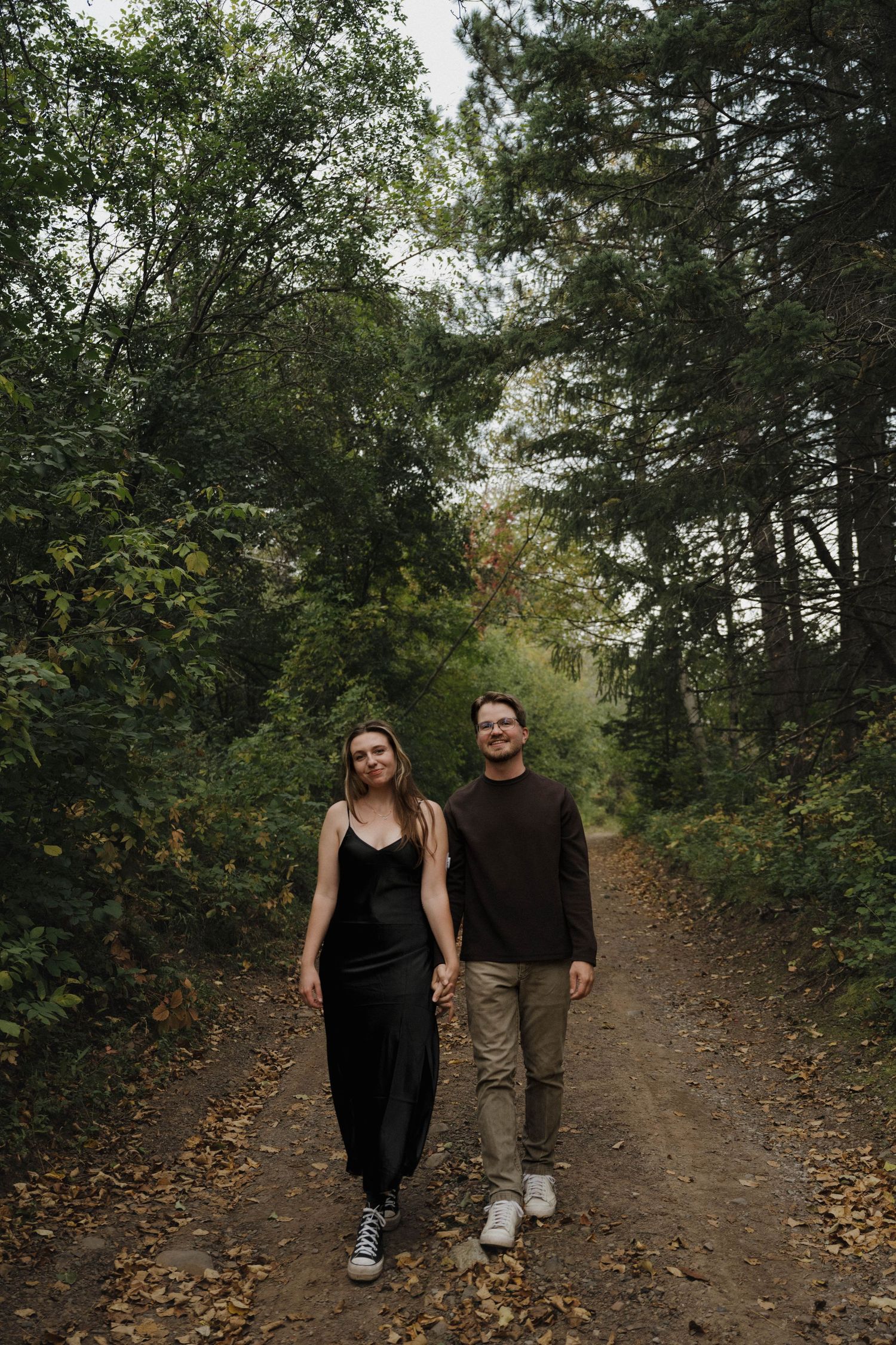 Couple holding hands walking on a forest dirt path, woman in black dress and man in brown sweater, surrounded by autumn trees.