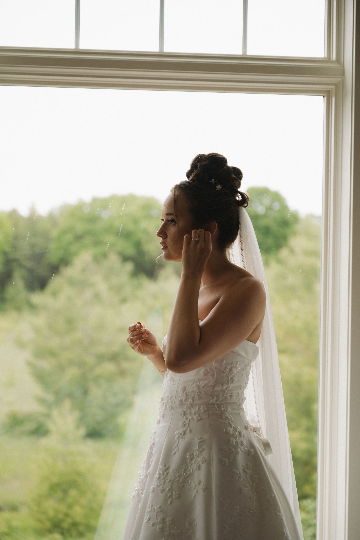 Elegant bride in lace gown and veil adjusting her earring while gazing out a large window.