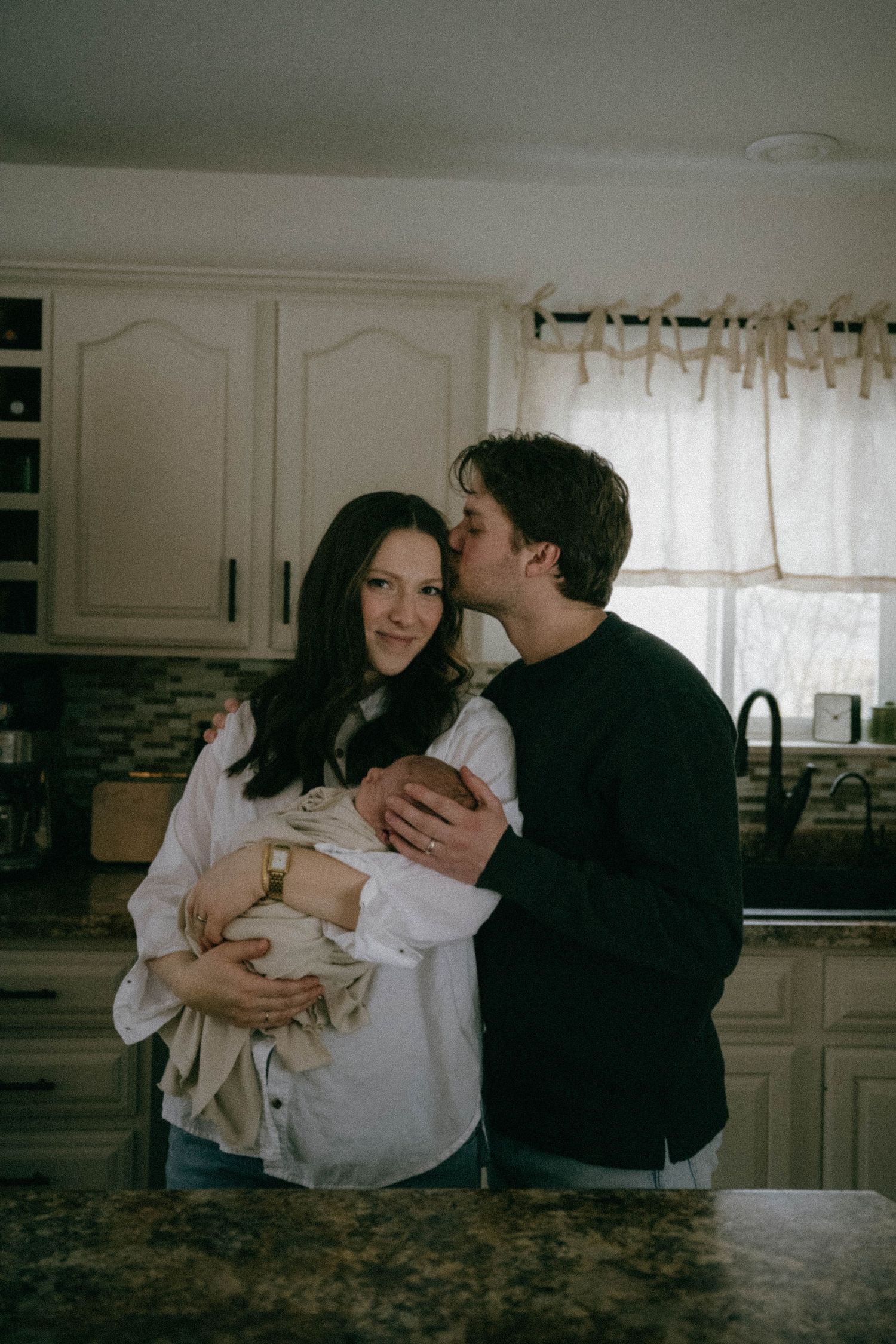 Happy couple stands in kitchen, man kisses woman's cheek as she holds newborn.