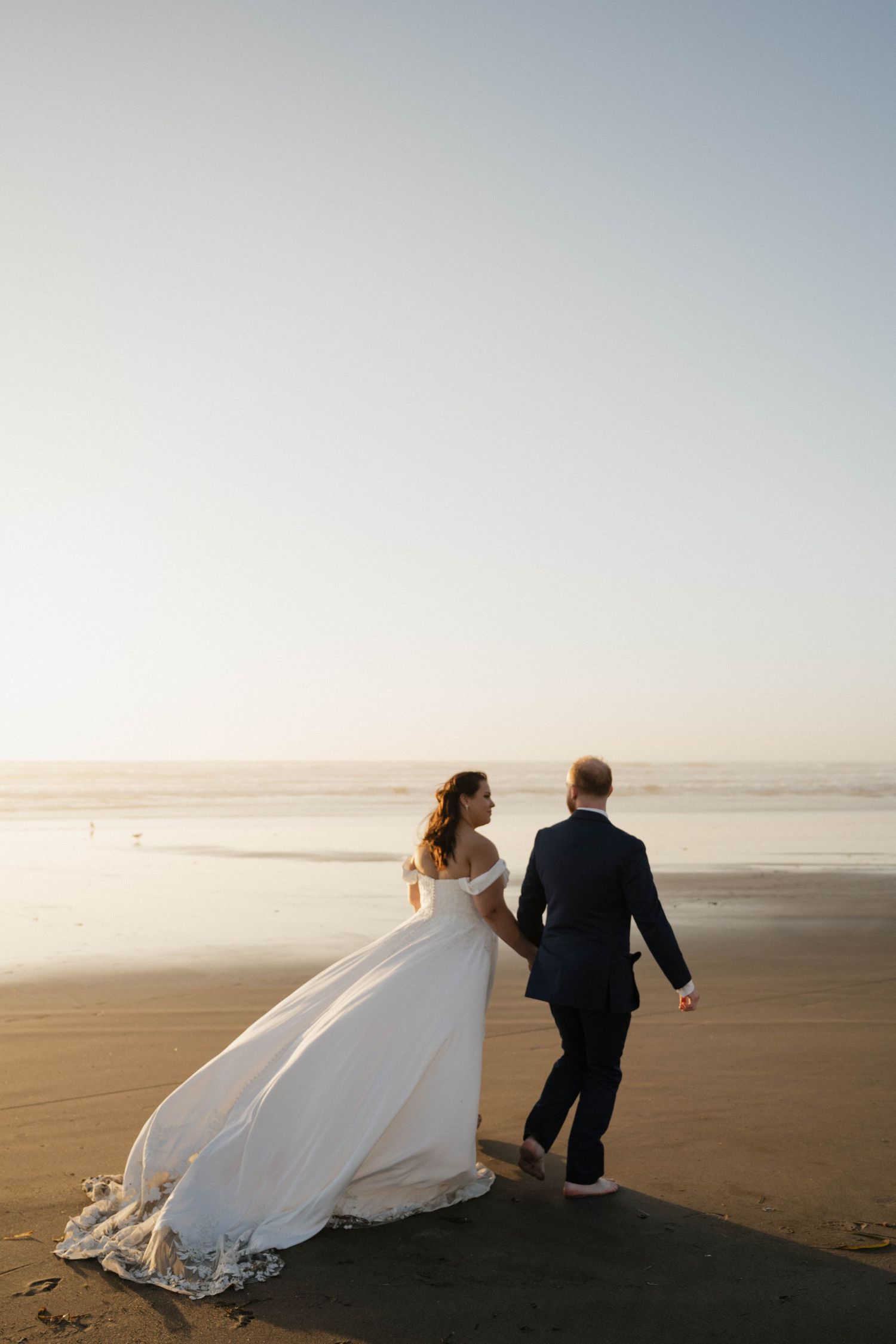 Bride and groom walk hand in hand along a misty beach at sunset, her white gown trailing behind.