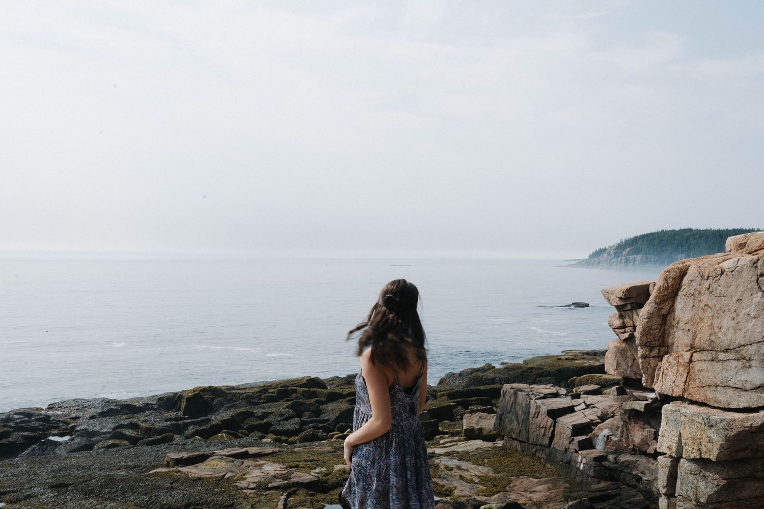 Woman in floral dress stands on rocky Maine coastline gazing at misty ocean view.
