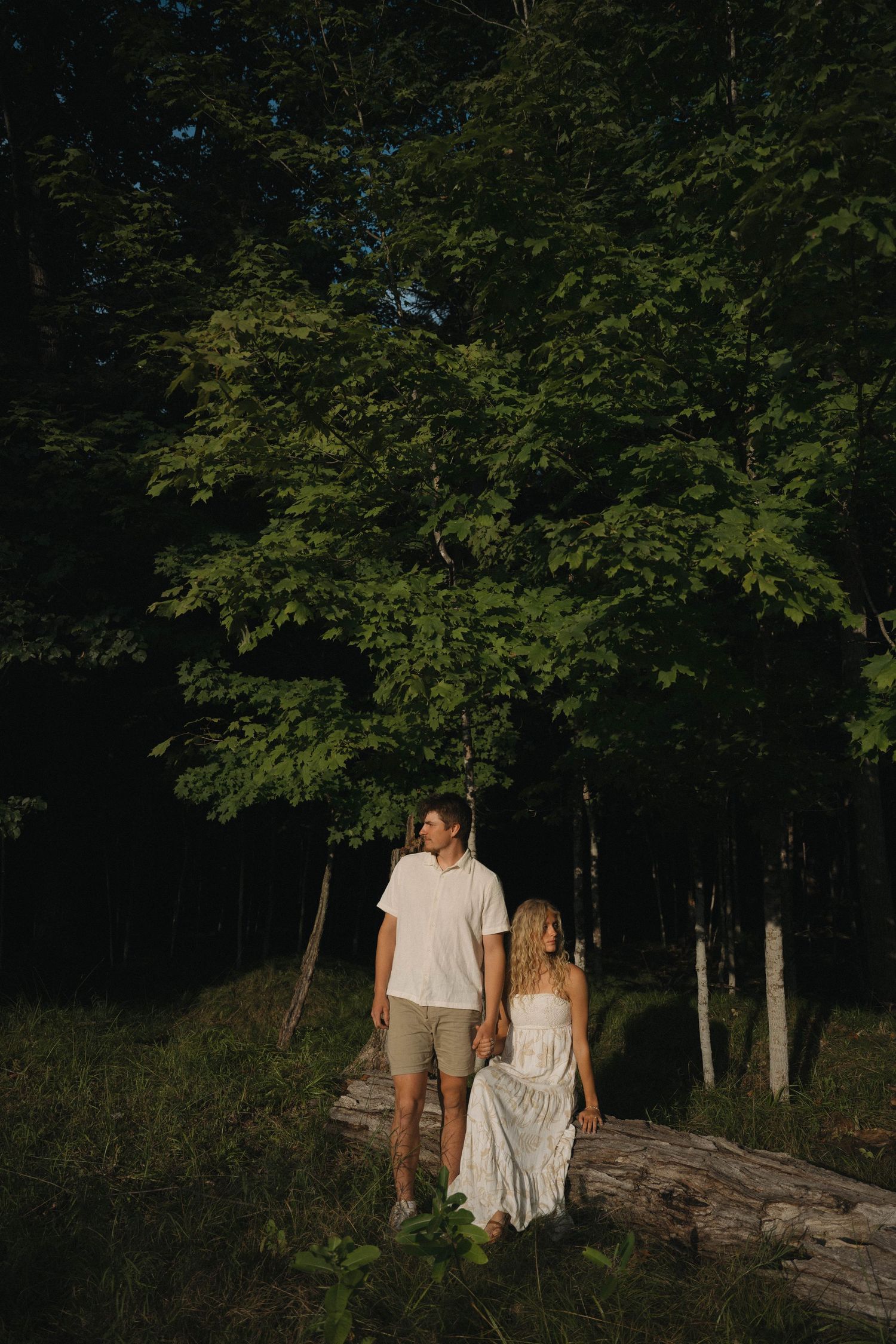 Couple standing together on a log surrounded by lush green forest trees.