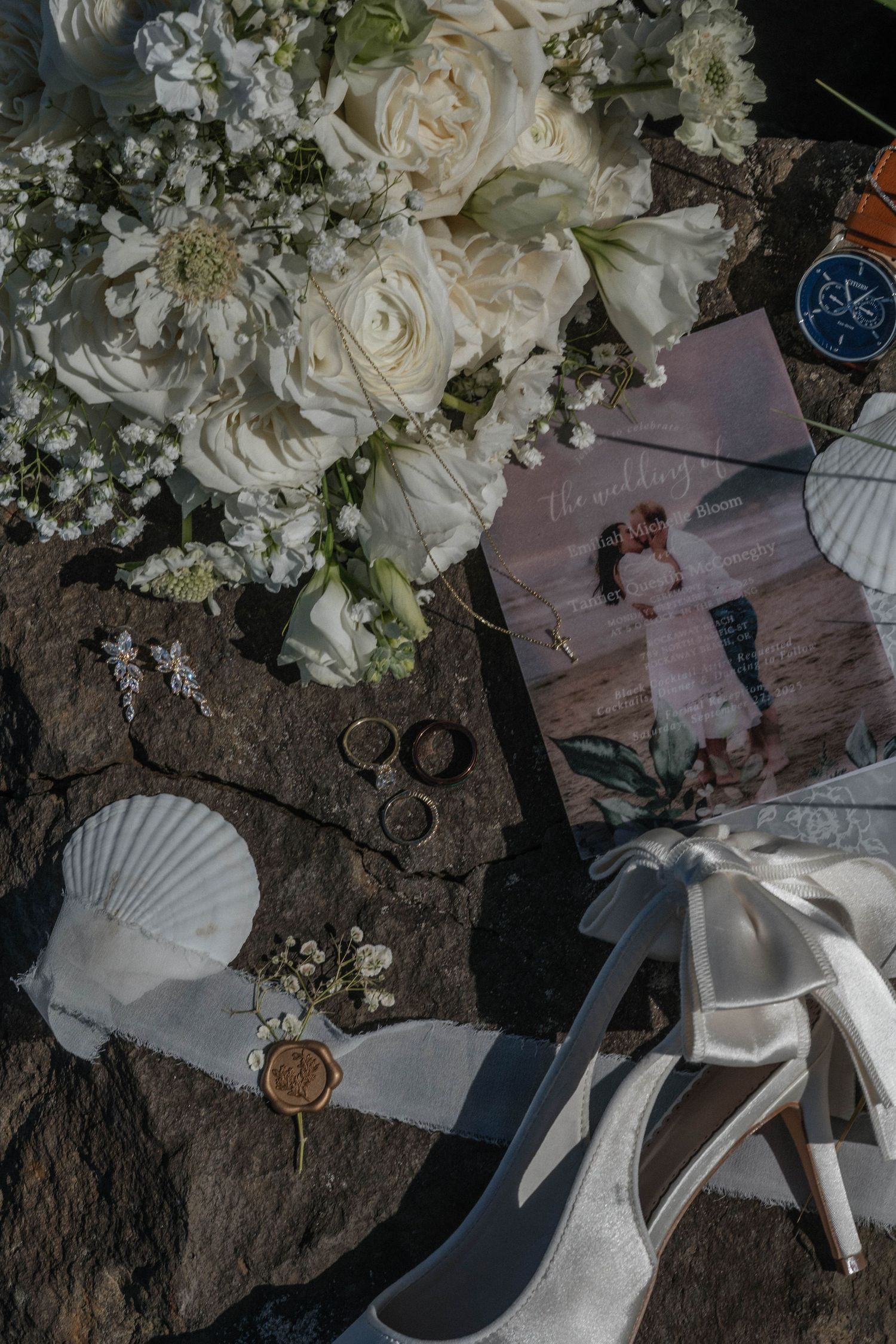 Flat lay of wedding details including white roses, rings, seashells, heels, and a wedding photo on rocks.