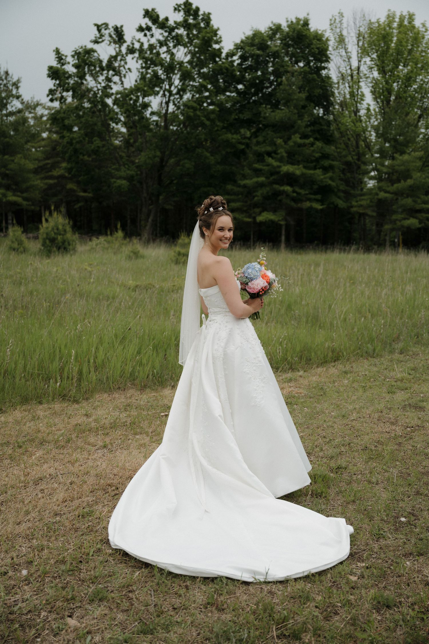 Bride in white gown with veil holds colorful bouquet in green field.