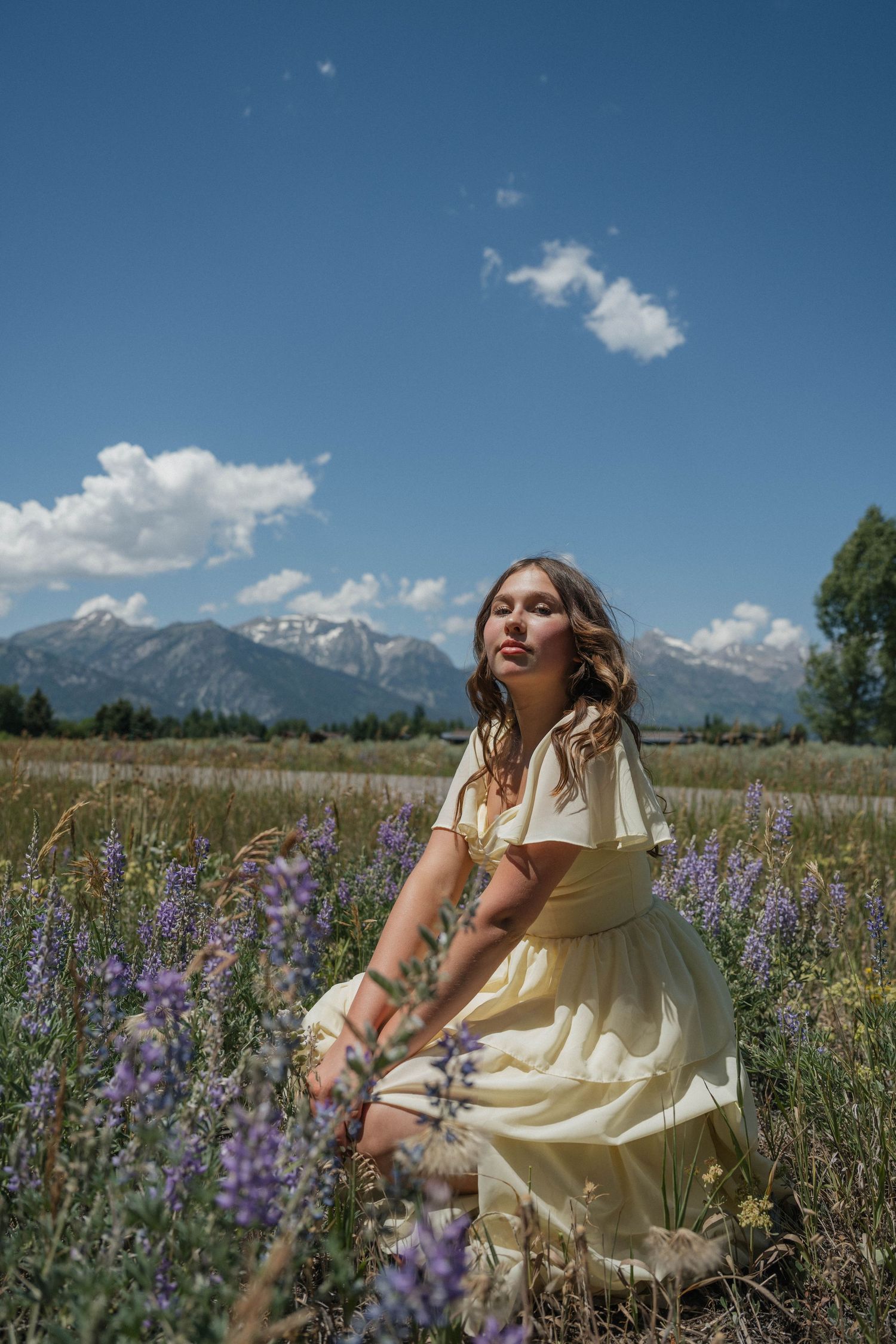 Young woman in yellow dress sitting among purple wildflowers with snow-capped mountains under blue sky.